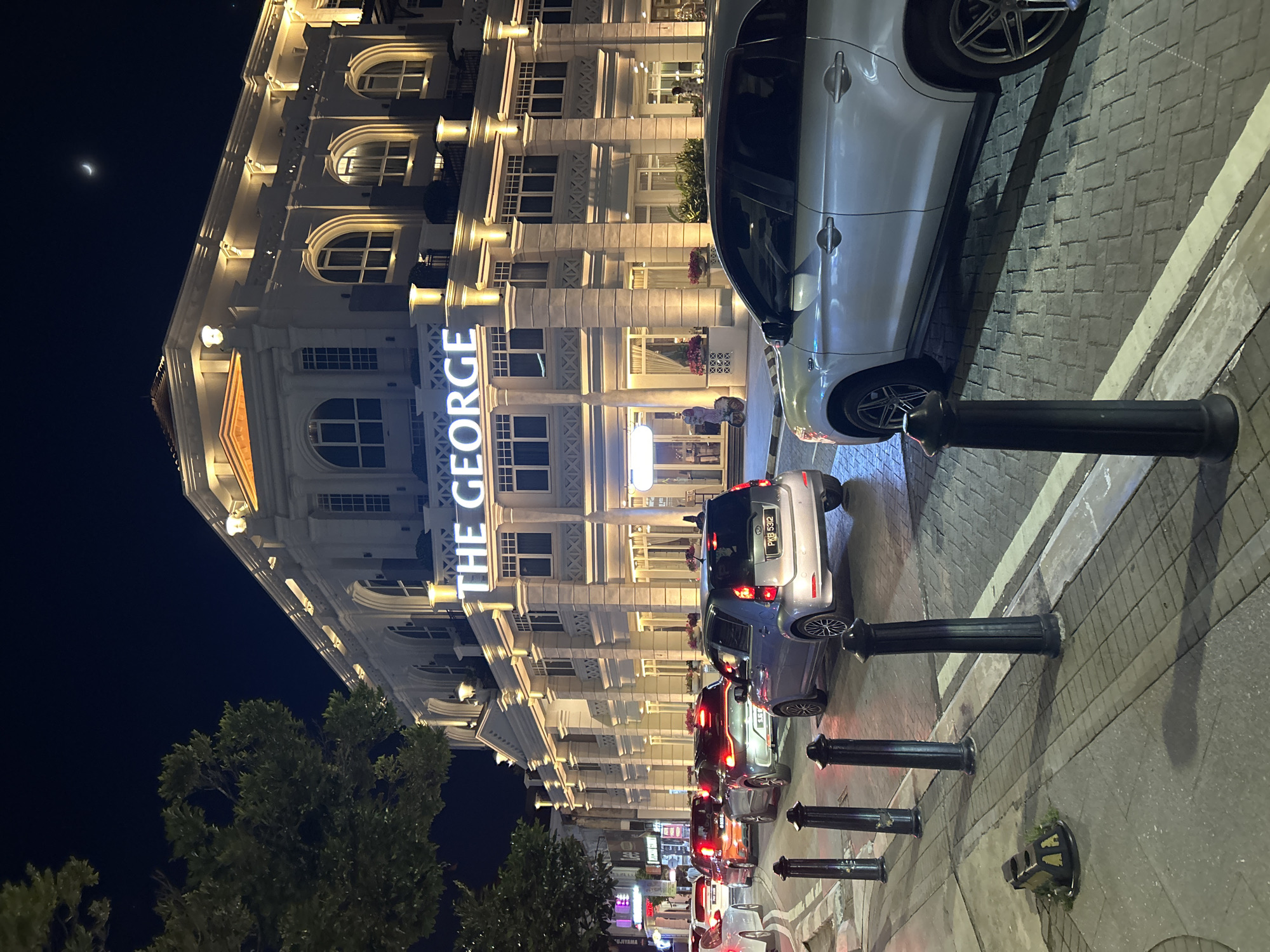 This is a nighttime, medium-shot, eye-level, vertical photograph of a street scene in George Town, Malaysia. In the foreground, a silver SUV is parked on the right side of the street, facing left. The vehicle is clean and appears to be in good condition. To the left of the SUV, a line of cars is driving on the road, their taillights illuminated. The road is paved with cobblestones, and a sidewalk is visible to the left of the road, lined with bollards.
In the background, a large, ornate building is illuminated by warm lighting. The building has multiple stories, arched windows, and decorative columns. A sign on the building reads "THE GEORGE" in large, white lettering. The sky above is dark, with a crescent moon visible in the upper right corner. A large tree with dark green leaves is present in the upper left quadrant of the image, partially obscuring the sky and building. The overall atmosphere of the image is one of urban nighttime activity.