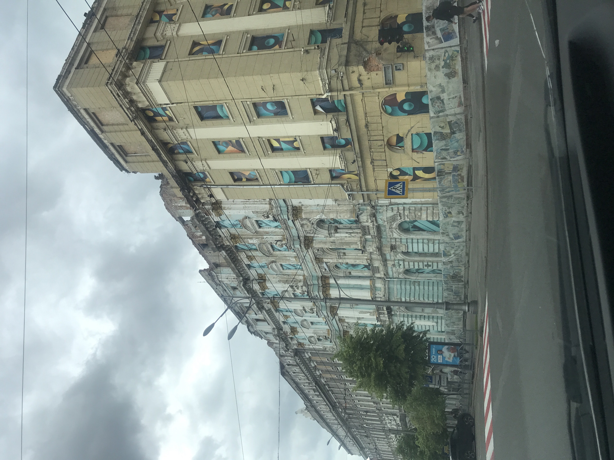 The image shows a street in Kharkiv, Ukraine, with two buildings of differing architectural styles. The first building is a pale yellow, multi-story structure with square windows filled with geometric artwork. On the ground floor, the facade features a line of arched openings, also decorated with artwork. A pedestrian crossing sign is visible on the building. The second building, located along the street, has a more ornate facade with detailed molding and architectural features painted in a muted blue and white color scheme. A row of trees lines the sidewalk in front of the second building. A street light stands in front of the trees. The sky is overcast with grey clouds. A portion of a vehicle's interior is visible in the right side of the image, suggesting the photo was taken from inside.