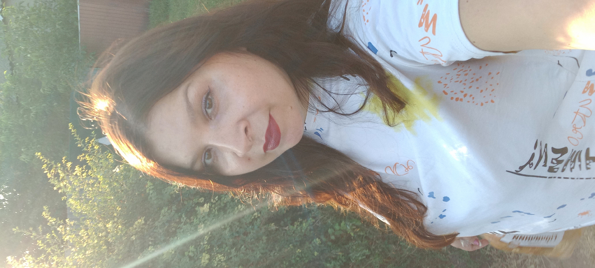 A medium shot, outdoor, waist-up portrait shows a woman with long, dark brown hair and dark red lipstick posing for a selfie. The sun is bright, casting a strong glare on her face and hair, and creating lens flare effects on the left side of the image. She is wearing a white t-shirt with an abstract pattern of orange, blue, and yellow splotches and lines. In the background, there are green bushes and trees, and a wooden fence. The image appears to be taken in a residential area on a sunny day. The location is identified as Klyntsi, Ukraine, though this context is not visually discernible from the image itself.
