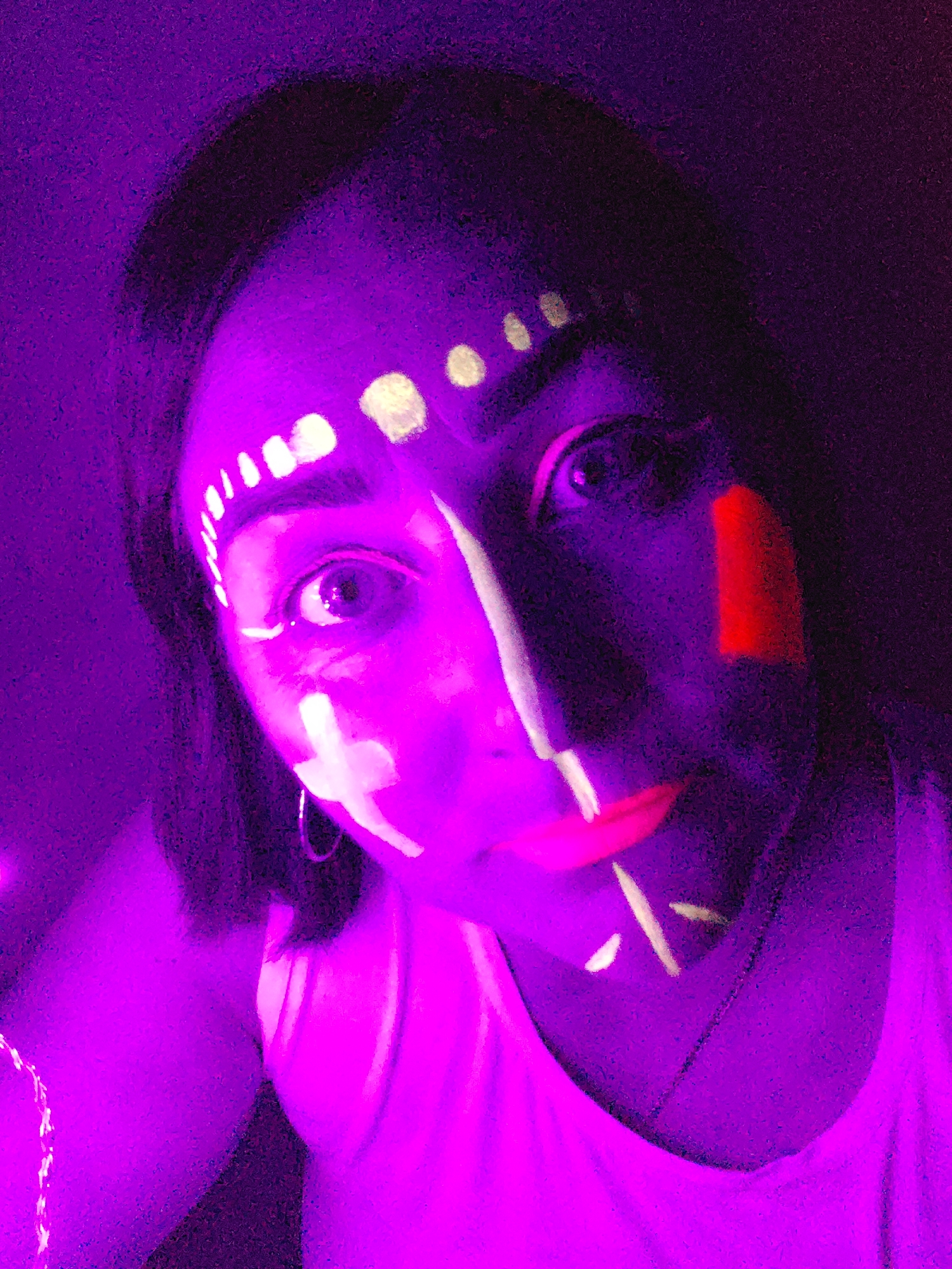 A person's face is illuminated with purple light and various white and orange markings. The markings include a row of dots on the forehead, a cross on the cheek, a vertical line down the nose, and short lines on the chin. There is a bright orange rectangle on the right cheek. The person has dark hair and is wearing a white tank top. A silver hoop earring is visible. The background is dark. There is no indication of the city of Markovskii, Russia in the image.
