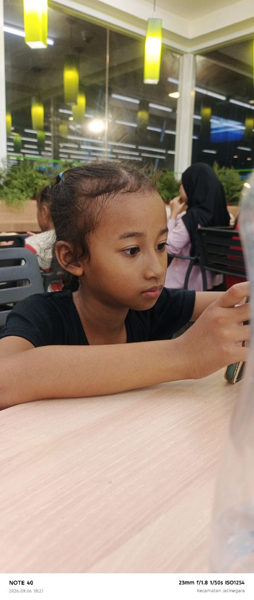 This is a vertical, close-up shot of a young girl sitting at a table. She has dark skin and dark, curly hair pulled back in a ponytail. Her eyes are cast downward, and her lips are slightly parted, suggesting she is focused on something out of frame. She is wearing a black t-shirt. Her arms are resting on the light-colored wooden table.
In the background, out of focus, are other people and elements of a dining setting. To the left, a blurry figure of another child is visible. To the right, a woman wearing a black hijab and a light pink top is seated at a table. The setting appears to be indoors, possibly a restaurant or cafe, with bright yellow, rectangular pendant lights hanging from the ceiling. The windows in the background show reflections and a dark interior with some green foliage.
The lower portion of the image contains text in a white font. In the bottom left corner, it reads "NOTE 40". Below that is the date "2026.09.06 18:21". In the bottom right corner, it reads "23mm f/1.8 1/50s ISO1254 Kecamatan Jatinegara". The date suggests the photo was taken on September 6, 2026, at 6:21 PM, indicating it is evening. The location "Kecamatan Jatinegara" places the scene in East Jakarta, Indonesia. The overall mood is one of quiet observation or engagement with a device.