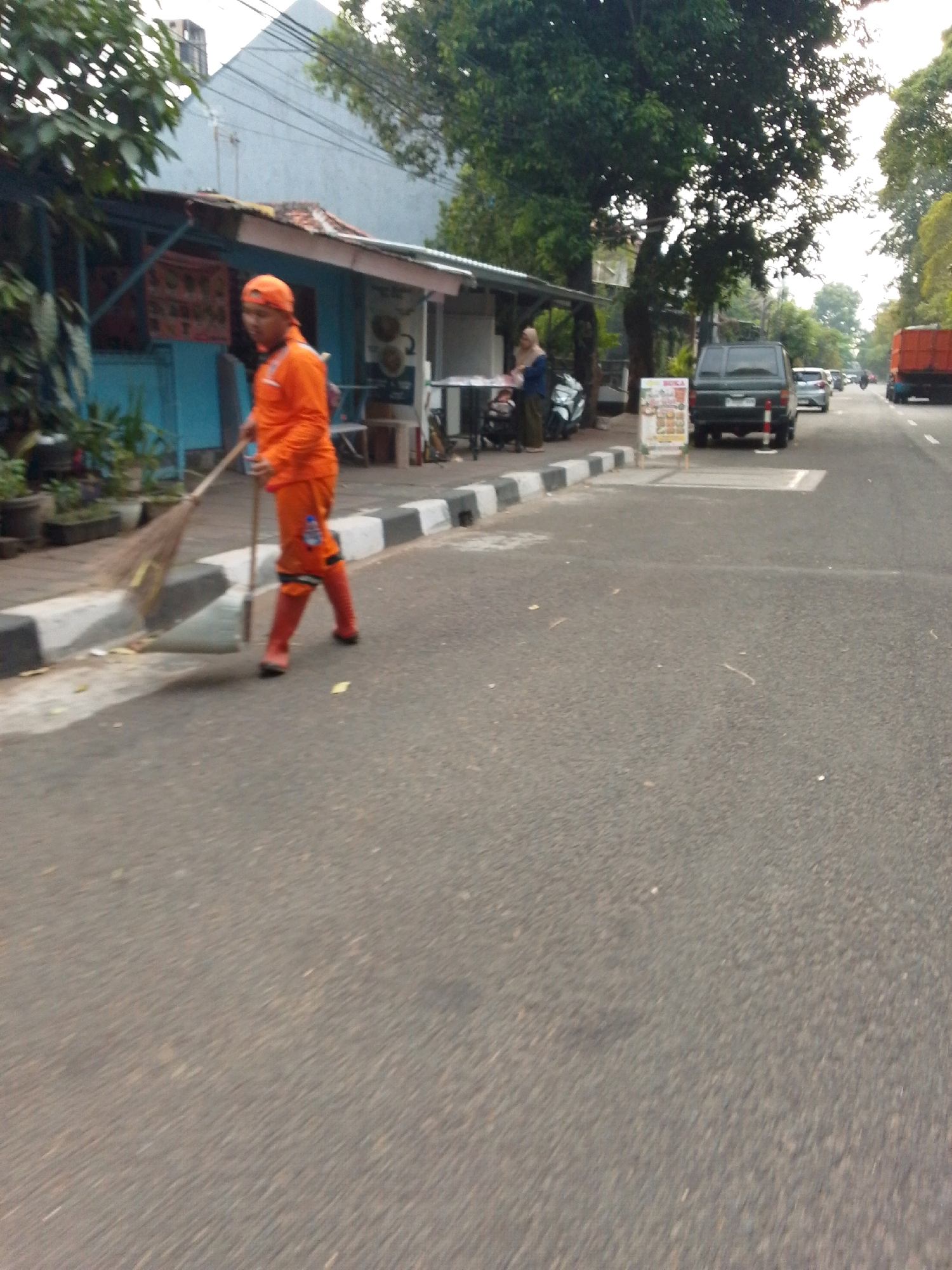 In East Jakarta, Indonesia, a sanitation worker in an orange uniform and cap is sweeping the side of a road. The worker is holding a broom and wearing orange boots, actively engaged in sweeping. To the left of the worker, a blue building with an orange-trimmed roof is visible, with potted plants in front. Behind the worker and the building, a large tree with dense foliage dominates the background. Further down the street, a dark-colored SUV and a red truck are present, along with other vehicles and pedestrians. The road is paved, and a concrete curb with black and white markings separates the road from the sidewalk.