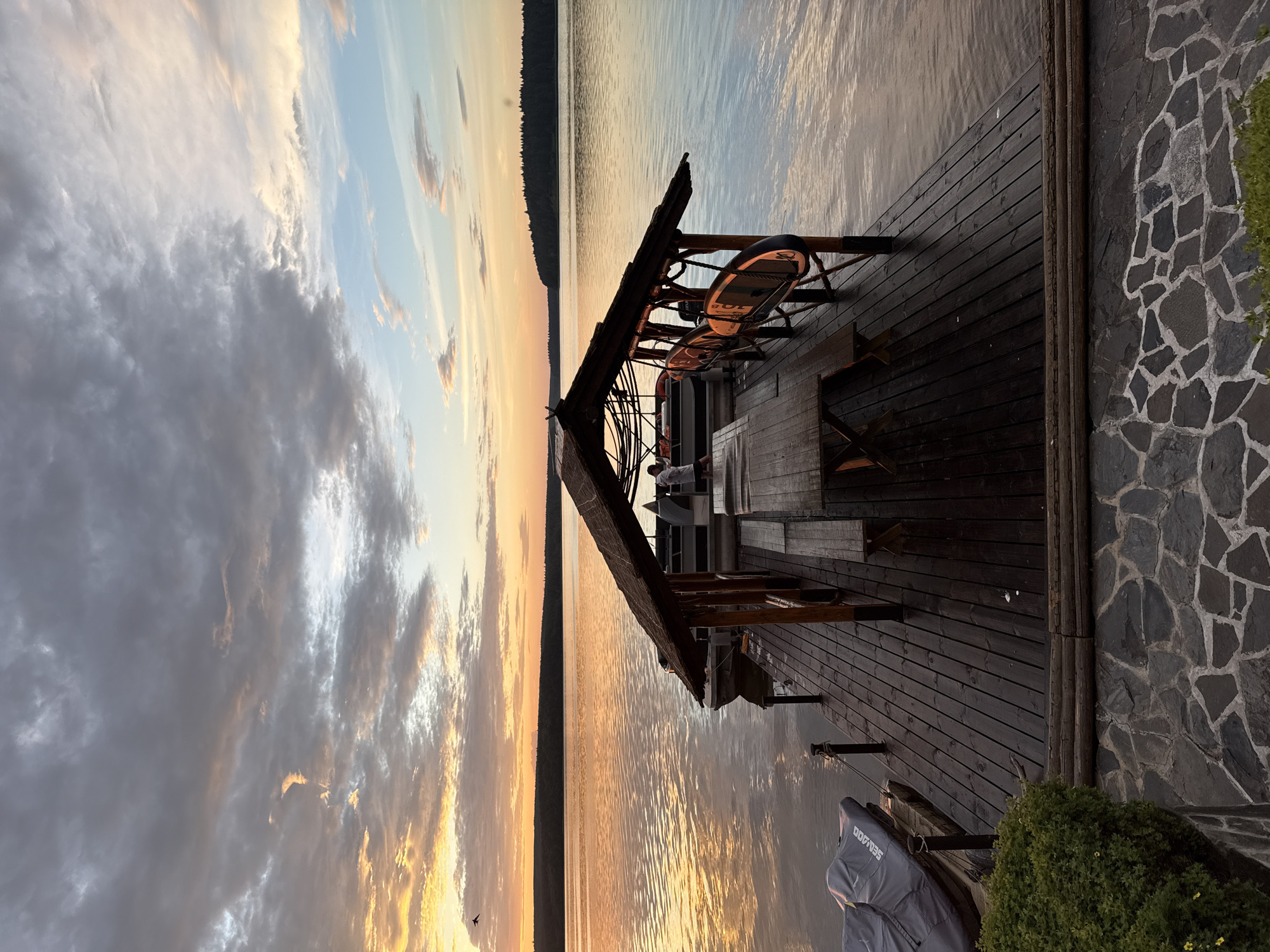 A wide shot captures a serene lakeside scene at sunset. The sky is a canvas of soft oranges, yellows, and blues, with dramatic clouds reflecting on the calm water. In the foreground, a wooden deck extends over the lake, featuring a covered seating area with tables and chairs. Two stand-up paddleboards are leaning against the structure, suggesting recreational activity. A person is partially visible sitting at one of the tables. The deck transitions to a stone-walled area on the right, bordered by a lush green bush. The overall atmosphere is tranquil and peaceful, indicating a relaxing evening by the water. The time of day is clearly sunset. The location is Votkinsk, Russia. Visible text includes "SEA-DOO" on a cover in the bottom right corner.