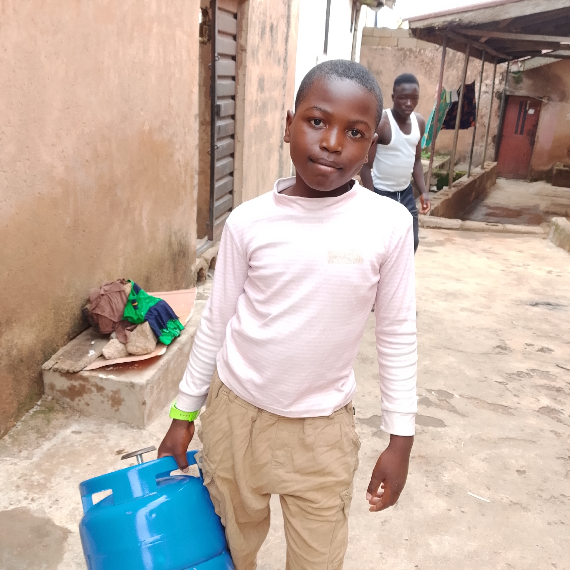 A young boy, likely of African descent, stands outdoors in what appears to be a densely populated urban area. He is positioned in the foreground, facing the camera and holding a bright blue gas cylinder. He is wearing a light pink, long-sleeved shirt with subtle horizontal stripes and tan cargo pants. A bright green wristband is visible on his left wrist.
In the background, another young man is visible, wearing a white tank top and dark pants. He appears to be walking away from the camera. The environment consists of a narrow pathway or alley between buildings with a rough, concrete ground. The buildings are constructed with weathered, light-colored walls and some corrugated metal roofing. Laundry is seen hanging on a line between two structures in the mid-ground. To the left of the boy, a pile of what appears to be bundled clothing and rocks is situated on a concrete ledge. The overall scene suggests a residential area with modest infrastructure. The time of day appears to be daytime, with overcast or diffused lighting.