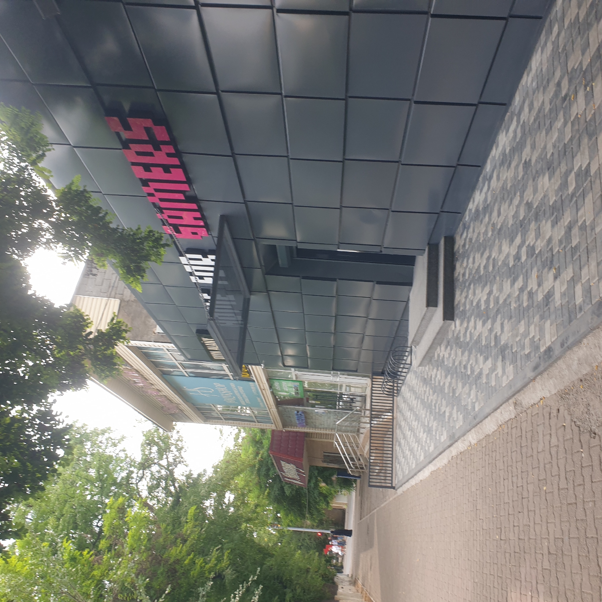 The image depicts a street scene in Shymkent, Kazakhstan. A modern building with a dark gray facade dominates the upper portion of the frame. Pink, block-style lettering on the building's facade reads "RAITHERS". Below the dark facade, there are glimpses of other businesses with signage, including what appears to be a "BOOKSHOP" with turquoise lettering and another with green signage.
The foreground shows a sidewalk paved with light brown interlocking bricks. Adjacent to this is a section of sidewalk paved with gray and white interlocking bricks, laid in a patterned fashion. A dark metal railing or fence is visible along the edge of the gray brick section.
Green trees with lush foliage line the street, some of which are close to the building, casting shadows on the facade and sidewalk. The sky, visible through the trees, is bright, suggesting it is daytime. The lighting indicates it's likely late morning or early afternoon with the sun relatively high in the sky, creating bright highlights.
In the lower part of the image, beyond the immediate storefronts, a street is visible with traffic lights and a few people walking. The overall scene suggests a commercial area on a clear day. The atmosphere appears calm and ordinary, with no specific event or activity clearly discernible beyond normal street life.