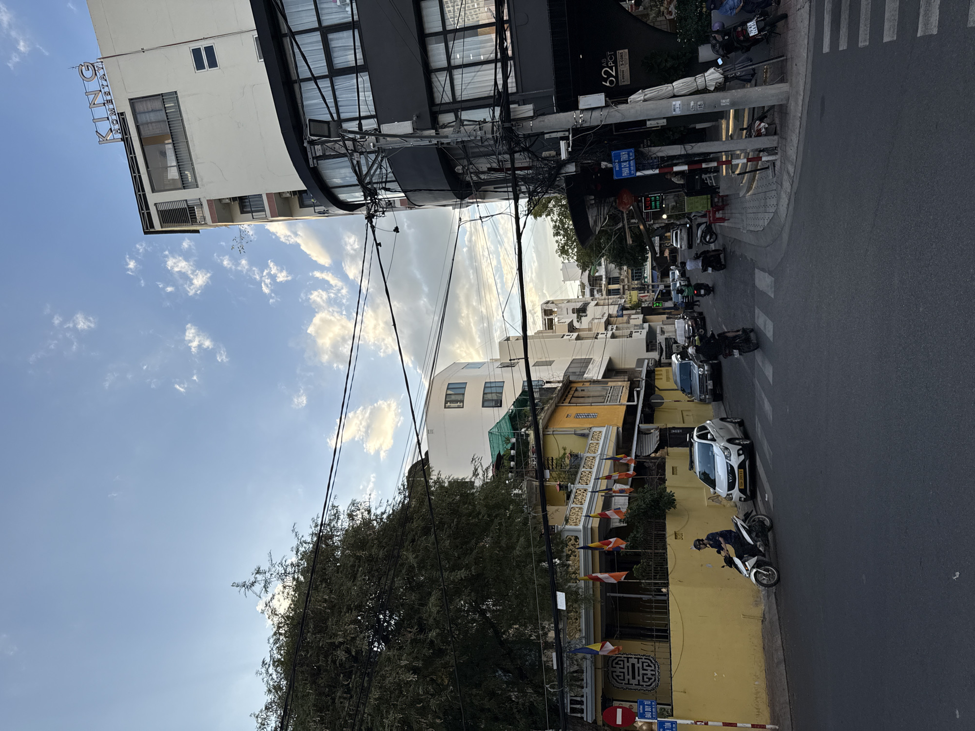 This image depicts a street scene in Nha Trang, Vietnam, likely taken in the late afternoon. A multi-story building with the sign "KING HOMESTAY" is visible on the right, featuring a curved, dark-colored facade with large windows. To its left, a yellow building with decorative railings and several flags is present. A large tree with dense foliage stands prominently on the left side of the frame. The street itself is paved and marked with a crosswalk. Several vehicles, including motorcycles and cars, are on the road. A person is riding a white scooter in the foreground, heading towards the left. Overhead, a network of electrical wires crisscrosses the sky, a common sight in Vietnamese cities. The sky is partly cloudy with a blue hue, suggesting a pleasant weather condition. The buildings indicate a mix of residential and commercial establishments.