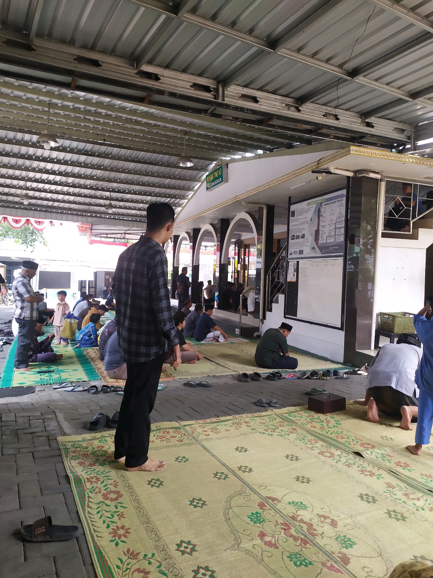 The image depicts an outdoor prayer area at a mosque in Sleman, Indonesia. Several men and boys are engaged in prayer or congregating on prayer mats spread across a brick floor. The prayer area is covered by a corrugated metal roof supported by a metal frame. To the right, a building with arched entrances and a white facade is visible. A large notice board is mounted on the building's exterior. The foreground shows a man in a black and white checkered shirt standing, facing away from the camera, with several pairs of sandals scattered on the ground. Other individuals are seen in various prayer postures, seated or kneeling on the prayer mats, which are adorned with floral patterns. The overall scene suggests a communal prayer gathering.