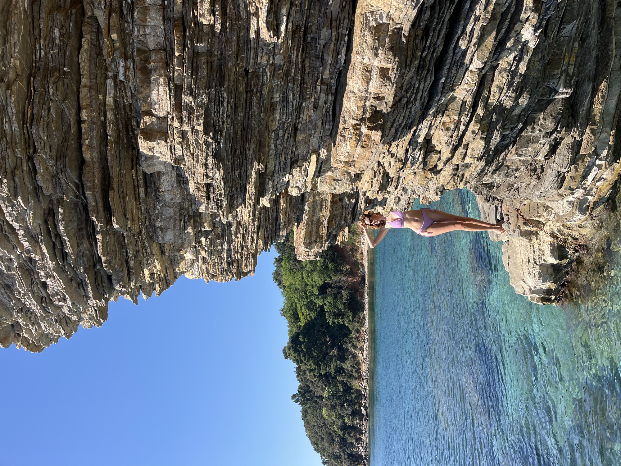 A vertical full shot shows a woman in a lilac bikini, standing on a rocky platform jutting out into turquoise water. She stands between two massive rock cliffs, one on the upper left and the other on the upper right, appearing to be part of the coastline. The water stretches out to a tree-lined shore in the distance, under a clear blue sky. The scene is likely in Budva, Montenegro, known for its dramatic coastline. The woman has her hands behind her head, suggesting a relaxed pose.