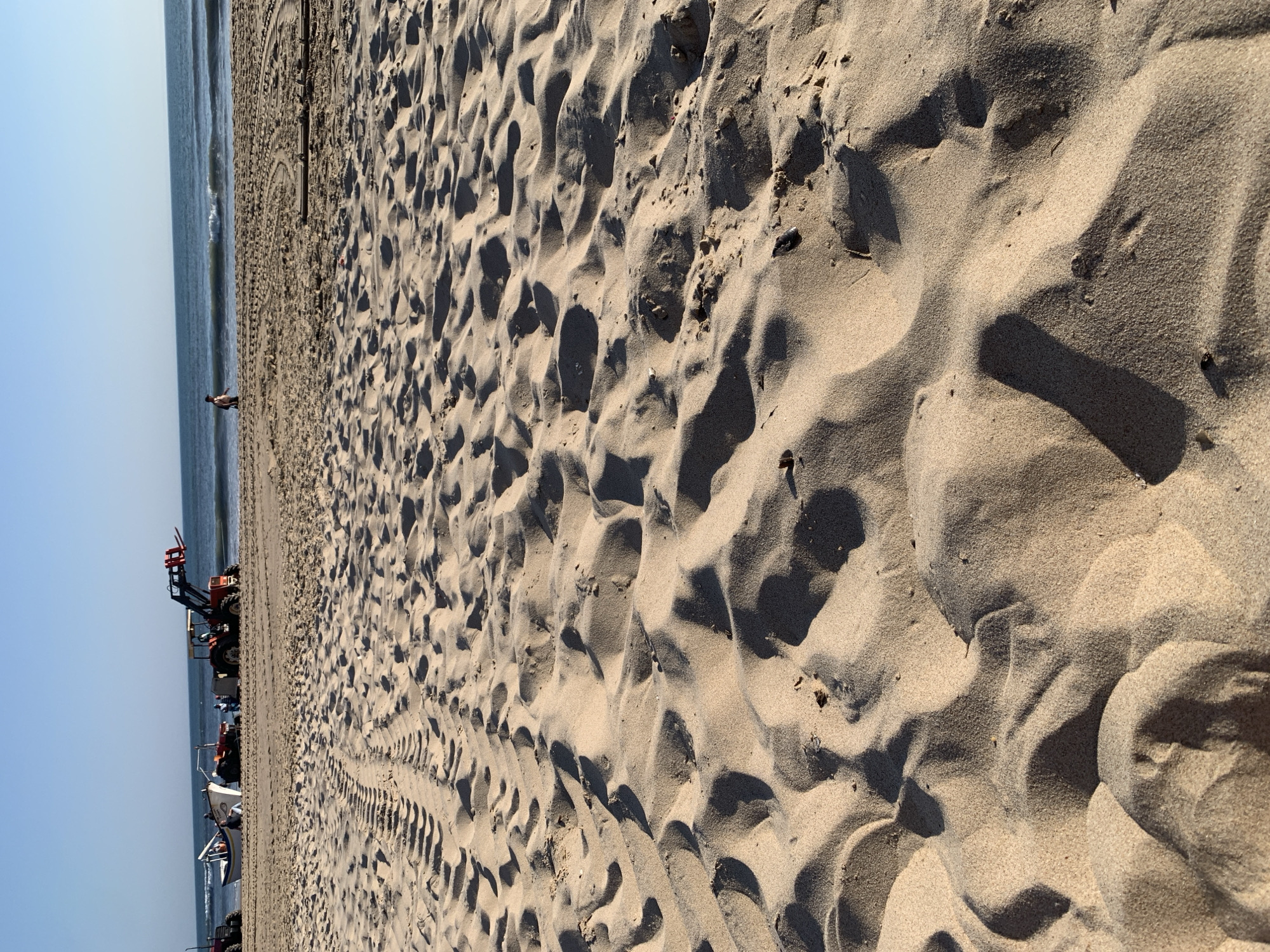 This image captures a wide view of a sandy beach in Costa da Caparica, Portugal, with the ocean in the background under a clear blue sky. In the middle ground, several tractors are positioned on the sand. One tractor, orange and yellow, has a loader attachment raised. Beside it, a white and blue boat is resting on the sand. Several people are visible near the boat and tractors, appearing to be working or engaged in activities related to the vehicles and boat. Further down the beach, a lone individual is walking near the water's edge. The foreground is dominated by the textured surface of the sand, showing ripples, tracks, and shadows cast by the sun. The scene suggests a working beach, likely involved in maritime activities such as launching or recovering boats.