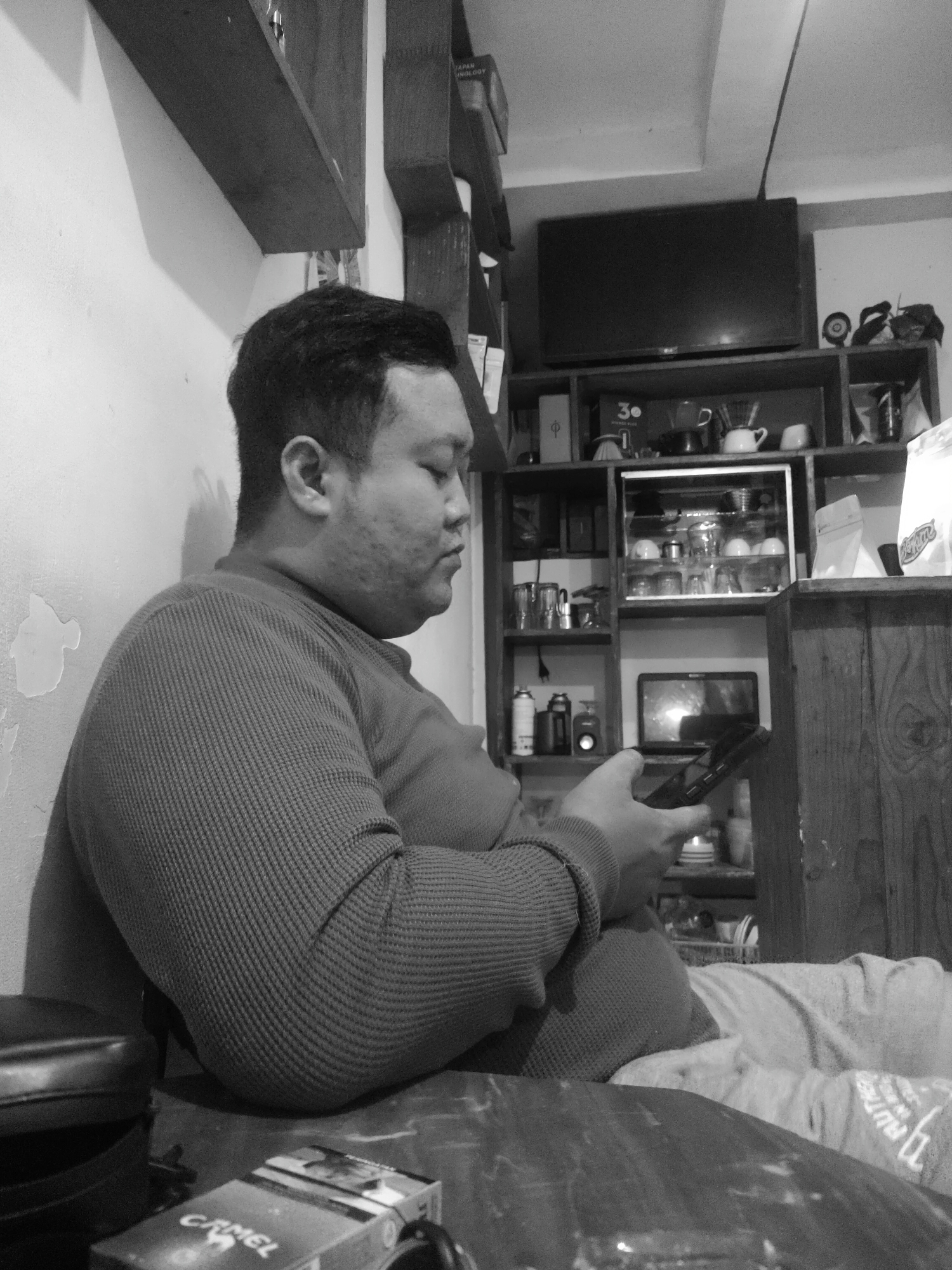 A black and white, medium shot photograph shows a man sitting against a wall, looking down at a phone in his hands. The man is wearing a long-sleeved, textured sweater. In the background, there is a shelf unit with various items including cups, a television, and bags. A pack of cigarettes is visible on the table in the foreground. The environment appears to be an indoor setting, possibly a cafe or residential space.