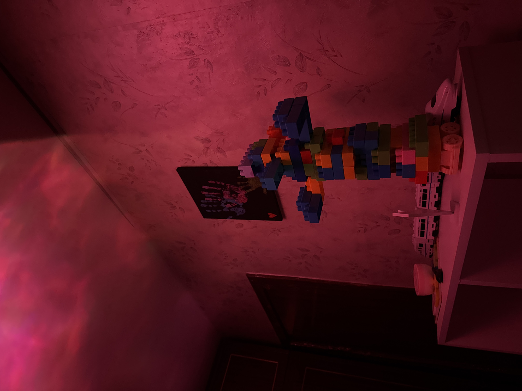 The image depicts an interior space with a red hue, possibly due to colored lighting. A colorful construction made of building blocks extends from a white shelf. A black rectangular object with colorful markings is visible near the construction. The walls have a patterned wallpaper. The setting is unknown. No people, actions, or interactions are visible. The location cannot be determined from the image.
