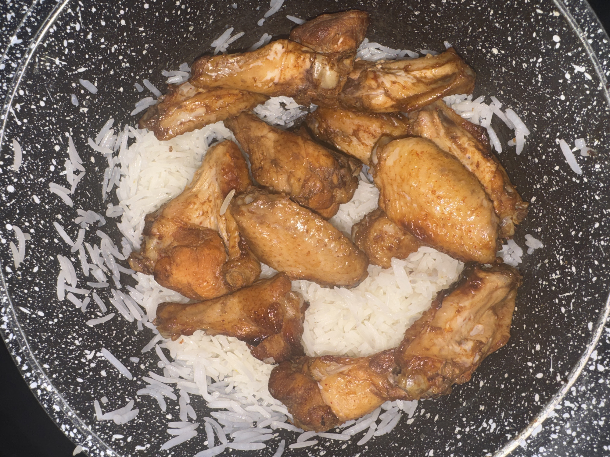 A close-up, overhead shot shows chicken wings coated in a dark glaze nestled in a bed of white rice. The food is contained within a speckled black frying pan. The scene is lit with overhead lighting, creating a somewhat harsh, shadow-filled image. There are no people present in the image, and the background is dark and indistinct.