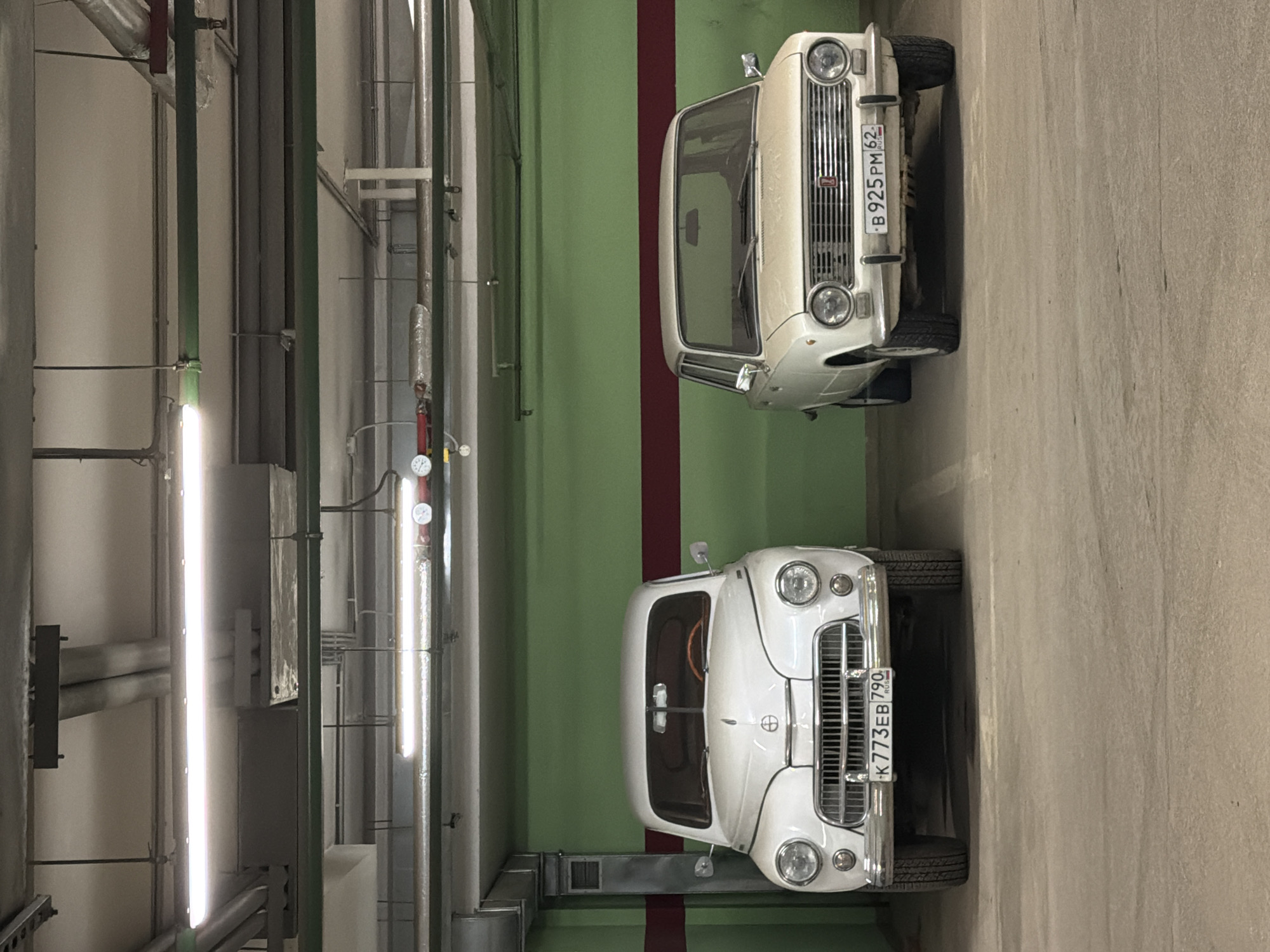 The image shows two vintage cars parked in an indoor parking space. The top car is a light-colored Lada parked adjacent to a wall painted with green and burgundy stripes. The license plate is partially visible: "B925PM62". The bottom car is a light-colored Moskvich with the license plate "K773EB790". The parking space has overhead pipes and lighting fixtures. The license plates indicate the cars are in Russia. The "62" region code on the Lada likely indicates the city of Ryazan, Russia.