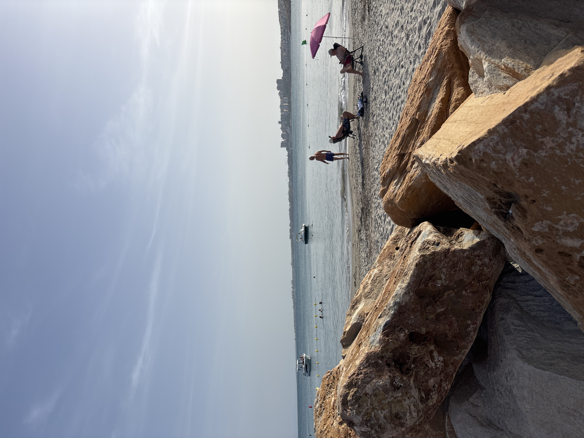 The image depicts a beach scene in the city of Orihuela, Spain. In the foreground, large, rough-textured rocks dominate the lower part of the frame. Beyond the rocks, a sandy beach stretches towards the sea. On the beach, three individuals are visible. Two men are seated in chairs, one under a pink umbrella, and the third man is standing by the water's edge. The sea is calm, with two boats anchored in the distance and several yellow buoys marking a roped-off area. The background features a distant cityscape with buildings and a clear, pale blue sky with wispy clouds. The lighting suggests it is late afternoon or early evening.