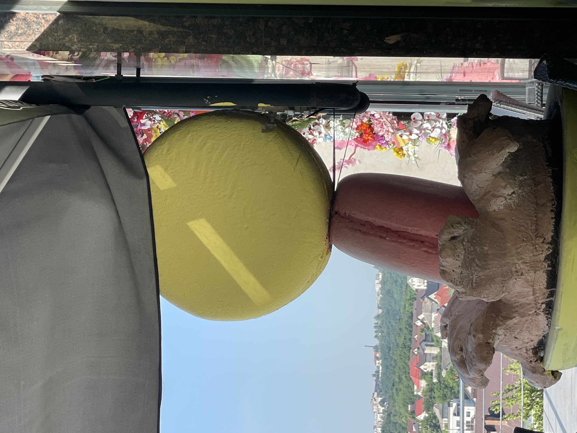 A large, yellow sphere, resembling a macaron shell, is balanced on top of a pink, elongated object, also resembling a macaron shell. The macaron sculptures are in the foreground, with a cityscape visible in the background under a clear blue sky. The cityscape features numerous buildings with red and white roofs, nestled among green trees. To the right of the macaron sculptures, there is a vertical black pole, and behind it, a wall with a textured, dark brown surface. In the mid-ground, a decorative arrangement of pink, red, and white flowers is visible, with some flowers appearing to be made of fabric. There is a hint of a grey awning or canopy overhead, casting a shadow. The overall impression is a whimsical, outdoor decorative display.