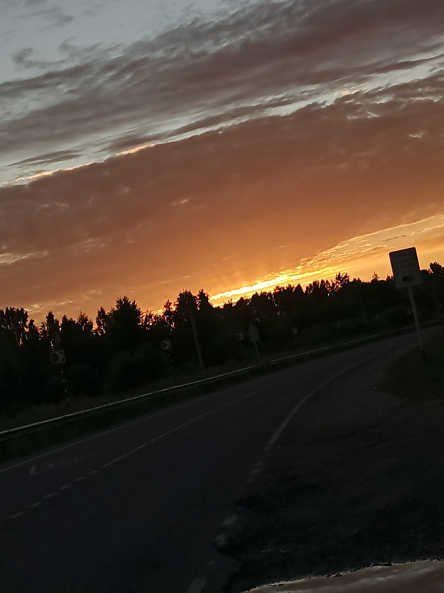 The image depicts a road at sunset near Voskresenskoye, Russia. The sky is a vibrant orange and yellow, with streaks of gray clouds running across it. A line of dark trees forms a silhouette against the colorful sky. A road, with white lane markings, curves through the foreground, and a guardrail runs along its edge. There are also traffic signs visible along the roadside. The overall scene is one of natural beauty during twilight.