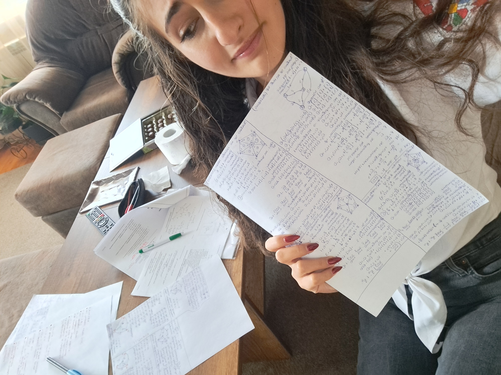 A young woman with long, dark, wavy hair is posing for a selfie indoors. She is holding a sheet of paper filled with handwritten mathematical equations and diagrams in front of her, obscuring part of her face. She is wearing a white shirt tied at the waist and dark gray jeans. Her fingernails are painted a dark red color.
On the wooden coffee table in front of her, there are several loose papers with similar mathematical content, a green pen, a blue pen, a calculator, a partially unwrapped box of chocolates, and a roll of toilet paper. In the background, there is a large brown couch. The location is unknown.