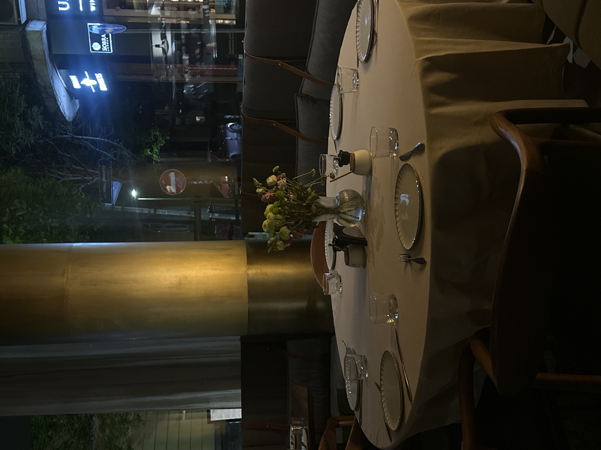 The image shows a partially set round table with a white tablecloth at a restaurant in Sofia, Bulgaria. The table is set with plates, glasses, cutlery, and a vase of flowers. There are chairs around the table, and a cushioned bench seat. In the background, through the restaurant window, the street outside is visible, including a building with illuminated signage.