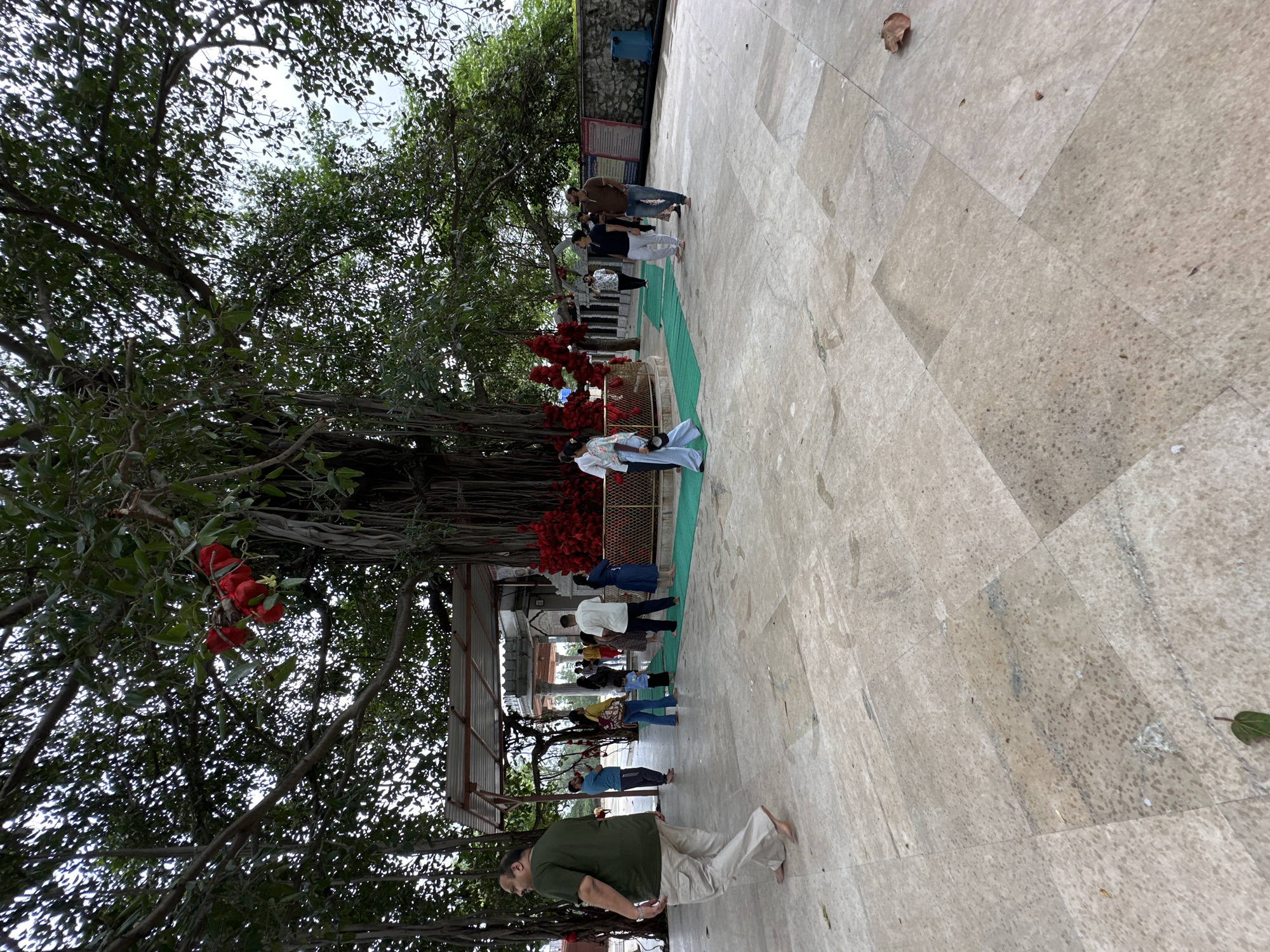 The image depicts a scene at a temple in Shahapur, India, likely under a large banyan tree. Several individuals are present, some walking, others standing, and some gathered around a structure. The ground is paved with large stone tiles. Red fabric decorations are visible around a central area, and the banyan tree's aerial roots are prominent. The environment appears to be outdoors, with a canopy of leaves from the tree overhead and a partially visible building structure in the background. Some individuals are dressed in casual attire, while others wear more traditional clothing. The overall atmosphere seems to be that of a public gathering place or a temple complex.