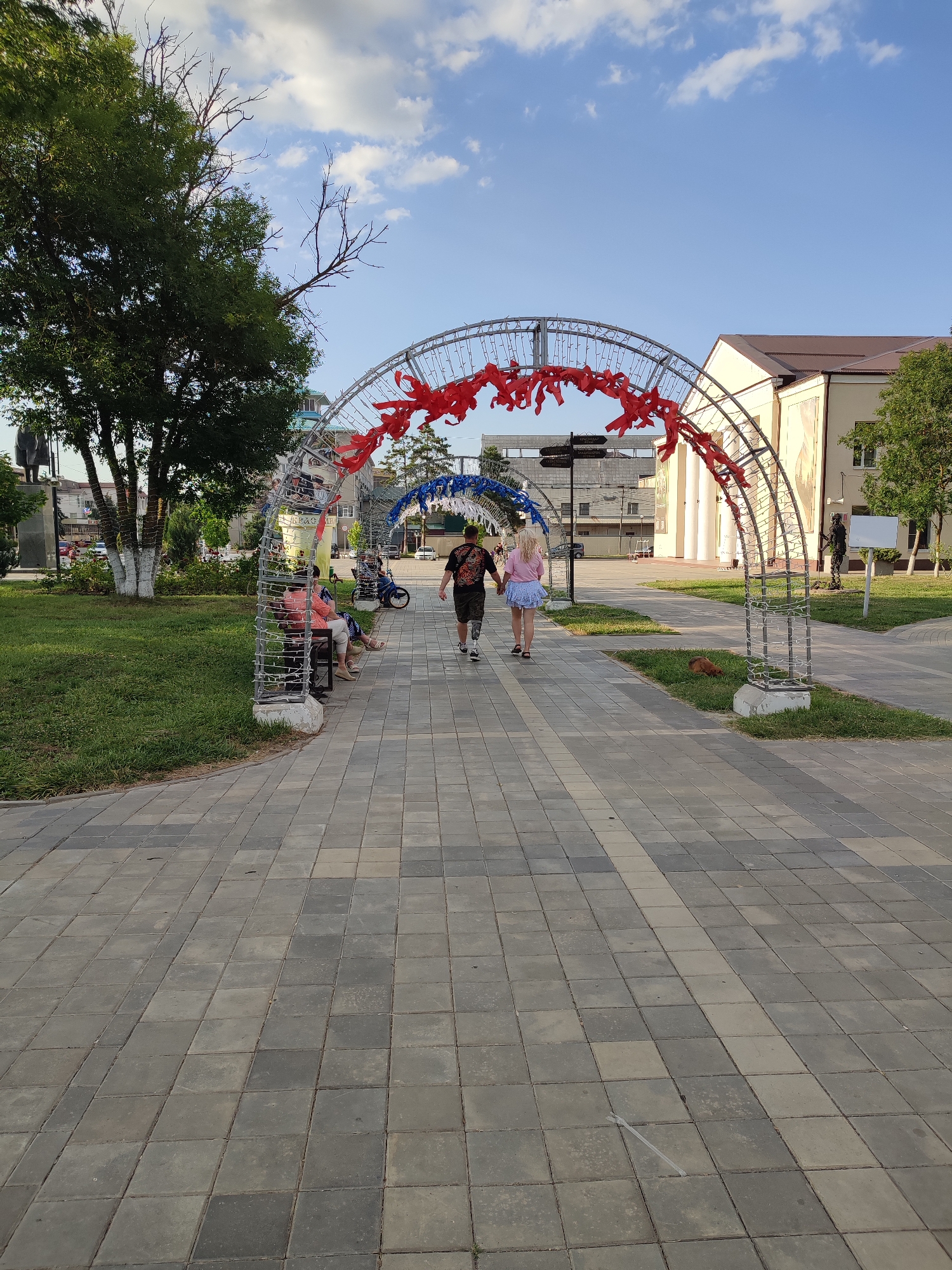 The image captures a paved walkway in a city park in Abinsk, Russia, on a clear day. Two young adults, a male and a female, walk away from the camera down the center of the walkway. The male wears a dark t-shirt with a graphic design and shorts, while the female wears a light pink t-shirt and a short, light blue skirt. They are walking under a large, arched metal structure decorated with red and white ribbons, which appears to be a festive archway. Further down the path, another similar archway, decorated with blue and white ribbons, is visible.
To the left of the walkway, a well-maintained green lawn is present, with a large tree and a statue of a figure in dark clothing. Several people are seated on a bench near the tree, including a woman in a pink top and a person in a white shirt. A bicycle is parked behind them.
To the right of the walkway, the grounds continue with patches of grass and trees. Behind the arches and lawn, a building with a brown roof and light-colored facade is visible, along with other structures and trees. The sky above is blue with scattered white clouds. The walkway is made of interlocking grey paving stones. A small, brown animal, possibly a dog or fox, is lying on the grass near the base of the archway.