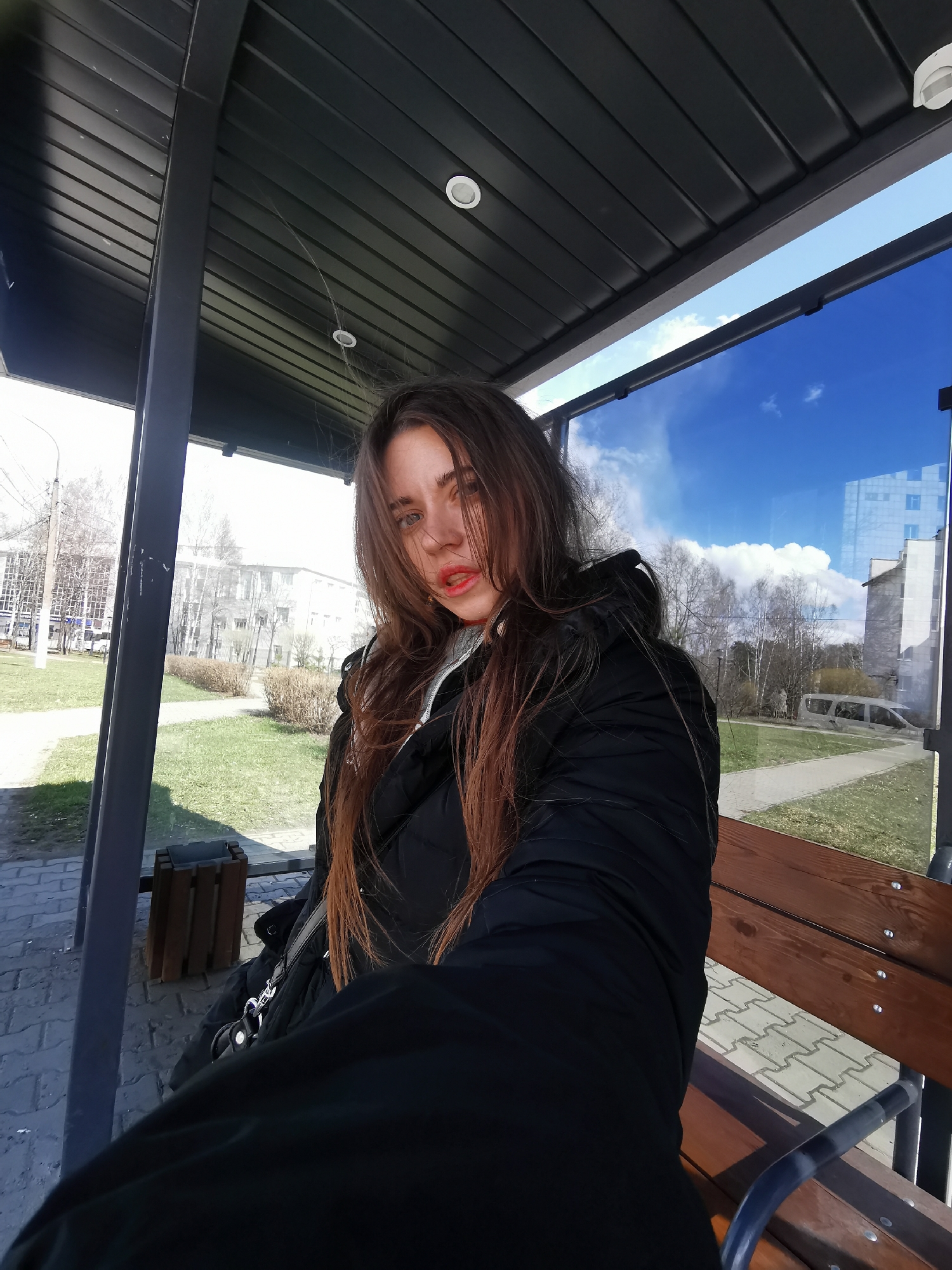 A young woman is depicted in a close-up, slightly low-angle portrait, sitting inside what appears to be a modern bus stop or a similar covered public shelter. She occupies the central-right portion of the frame, looking directly at the camera with a neutral, unsmiling expression. Her long, dark brown hair is prominent, framing her face and appearing somewhat disheveled or wind-blown. She has light-colored eyes and is wearing vibrant red or orange lipstick.
She is dressed in a black, puffer-style jacket or coat, and a strap from a dark-colored bag is visible over her left shoulder. She is seated on a wooden slatted bench with dark metal supports.
Above her, the shelter's ceiling is dark grey with a ribbed or slatted design, featuring two circular recessed lights. The structure also includes prominent dark grey vertical beams.
The background, visible through the glass panels of the shelter and reflecting the environment, shows a bright, sunny day. A clear blue sky with fluffy white clouds dominates the upper background. Below, an urban or semi-urban landscape unfolds, featuring areas of green grass, some patches of drier vegetation, and several bare trees, suggesting late winter or early spring. Multi-story buildings are visible in the distance, and paved pedestrian areas run along the ground. A light-colored vehicle is faintly reflected in the glass on the right. A wooden slatted trash bin is also visible near the ground on the left.
The time of day appears to be bright daylight, likely mid-morning or early afternoon, given the clear skies and strong illumination. The overall scene suggests a cool but sunny day. No discernible text is visible in the image.