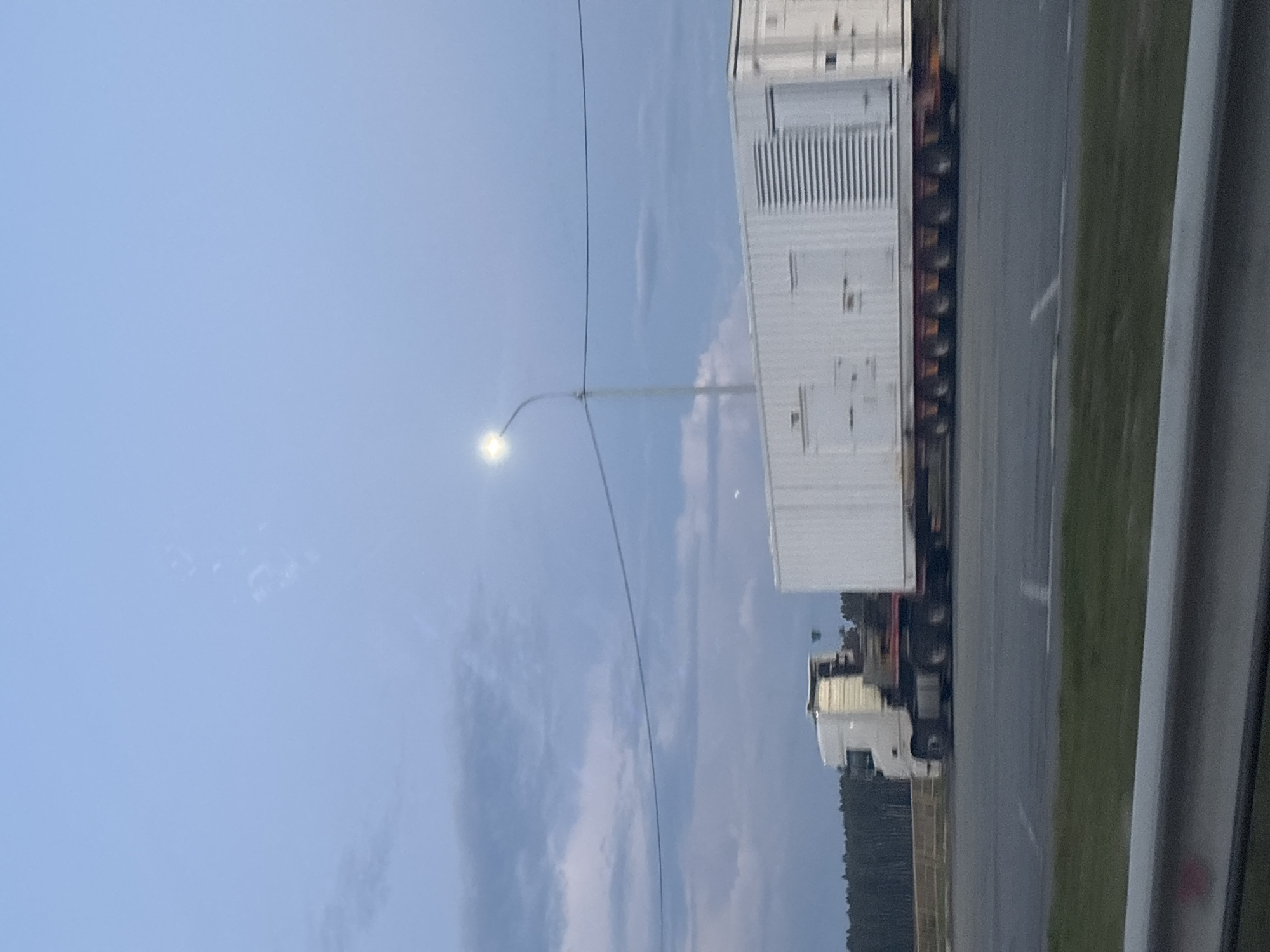 A white semi-truck transports a large white container with ribbed panels and doors down a paved road with a line of trees in the background. The sky is a hazy blue with some scattered clouds, and a streetlamp is visible.