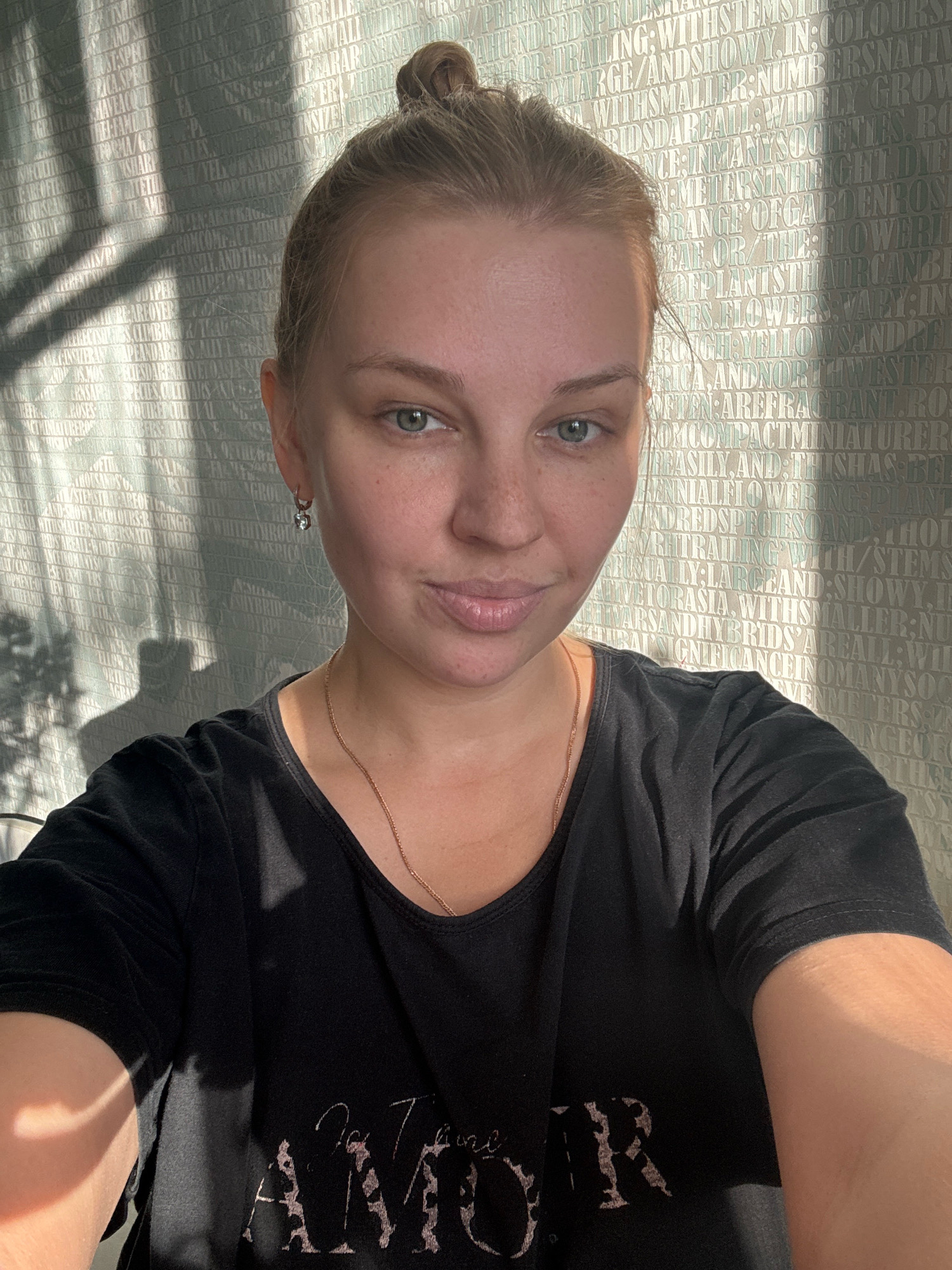 A young woman with blonde hair pulled into a bun is seen in a close-up selfie. She is wearing a black t-shirt with a gold necklace. The background is a patterned wallpaper with text. Sunlight casts shadows across her face and the background. There is no indication of the location being Rusanovo, Russia.