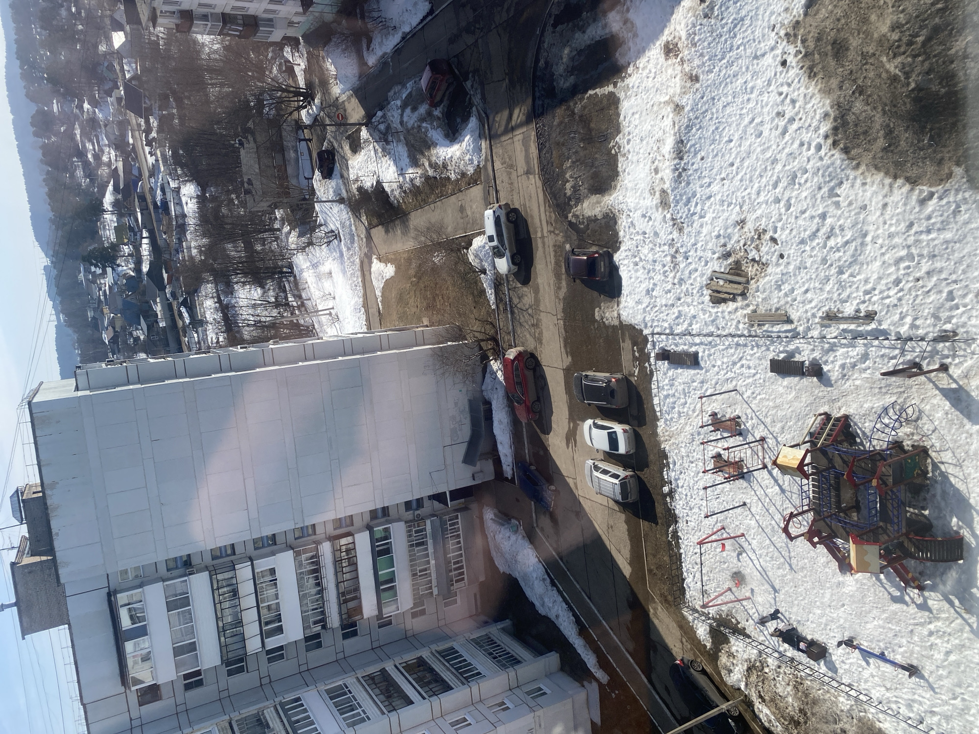 This elevated image provides a forensic view of a residential area in Chaikovsky, Russia, during late winter or early spring, characterized by melting snow and bare vegetation under a clear, bright sky.
The immediate foreground features the lower levels of a tall, light-grey multi-story panel apartment building with numerous windows and enclosed balconies. Adjacent to this structure is a paved parking area and access road, partially covered by snow and slush. Five vehicles are parked parallel to the building: a red SUV, a blue sedan, a white SUV, a light grey SUV, and a dark grey sedan. A white SUV is positioned on a connecting paved road, near an intersection marked by a "STOP" sign. A dark red car is parked further down this road.
A snow-covered children's playground occupies the bottom portion of the scene. It includes a prominent multi-level climbing structure with slides, stairs, and tunnels, primarily colored in red, blue, and yellow. Two sets of swings with red frames and a blue-and-red seesaw are also present. A horizontal ladder structure is visible on the left side of the playground. Two individuals are within the playground area: one person in light outerwear walks near the horizontal ladder, and another person is seated on a bench nearby. Footprints are discernible in the melting snow.
Beyond the immediate building and playground, the midground consists of a mix of paved roads and unpaved terrain with patches of exposed brown earth and melting snow. Numerous bare trees and sparse shrubs are scattered throughout. The background is dominated by a forested hill range under a clear blue sky, featuring dormant deciduous trees and scattered residential structures on its lower slopes. Utility poles and overhead lines are visible traversing the midground.