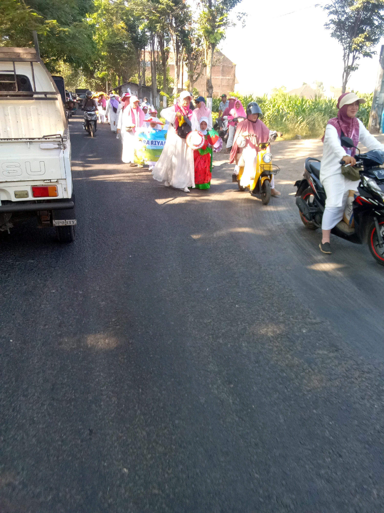 The image depicts a street scene in Pancakarya, Indonesia, under bright sunlight. A procession is underway, featuring several individuals. In the foreground left, a white truck with the visible text "SU" on its side is parked. A line of people are walking along the road. The individuals are dressed in traditional attire. Some are wearing white robes and headscarves, while others are adorned in colorful clothing.
Several people are riding motorcycles and scooters. One person on a yellow scooter wears a pink headscarf and a pink cloak. Another individual on a black motorcycle wears a white outfit and a maroon headscarf. A child in a red costume with green trim is visible amongst the walkers, holding a banner that says "PA RIYA." The road is paved and appears to be an urban or semi-urban street, with trees lining one side of the road and buildings in the background. The overall atmosphere suggests a celebratory or cultural event.