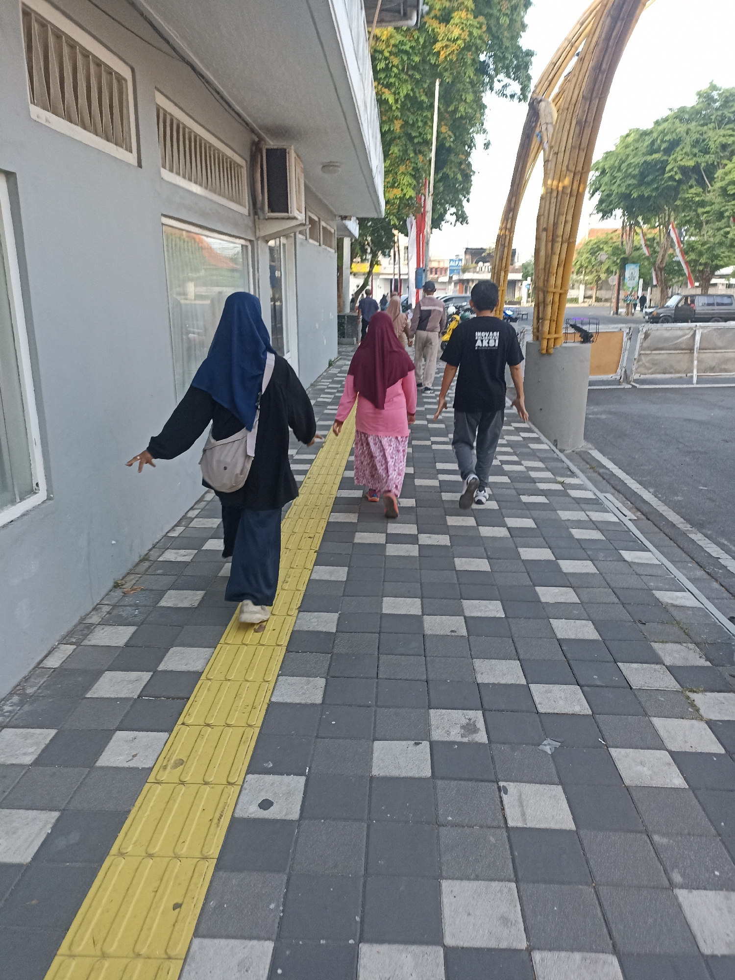 In Kelurahan Kepatihan, Indonesia, this image captures a street scene with a sidewalk patterned with gray and white square tiles, bordered by a prominent yellow tactile paving strip. Three individuals are walking away from the camera along the sidewalk. The first person, on the left, wears a dark blue hijab and a dark jacket over blue pants, carrying a beige crossbody bag. The second person, in the middle, is wearing a maroon hijab and a light pink patterned long-sleeved shirt with a long skirt. The third person, on the right, is a male wearing a black t-shirt with white text that reads "INOVASI AKSI" and grey pants. They are walking towards a large, arched bamboo structure. In the background, trees, buildings, a street with vehicles, and a pedestrian crossing are visible, suggesting a bustling urban environment. The lighting indicates it is daytime, possibly late afternoon due to the soft light and shadows.
