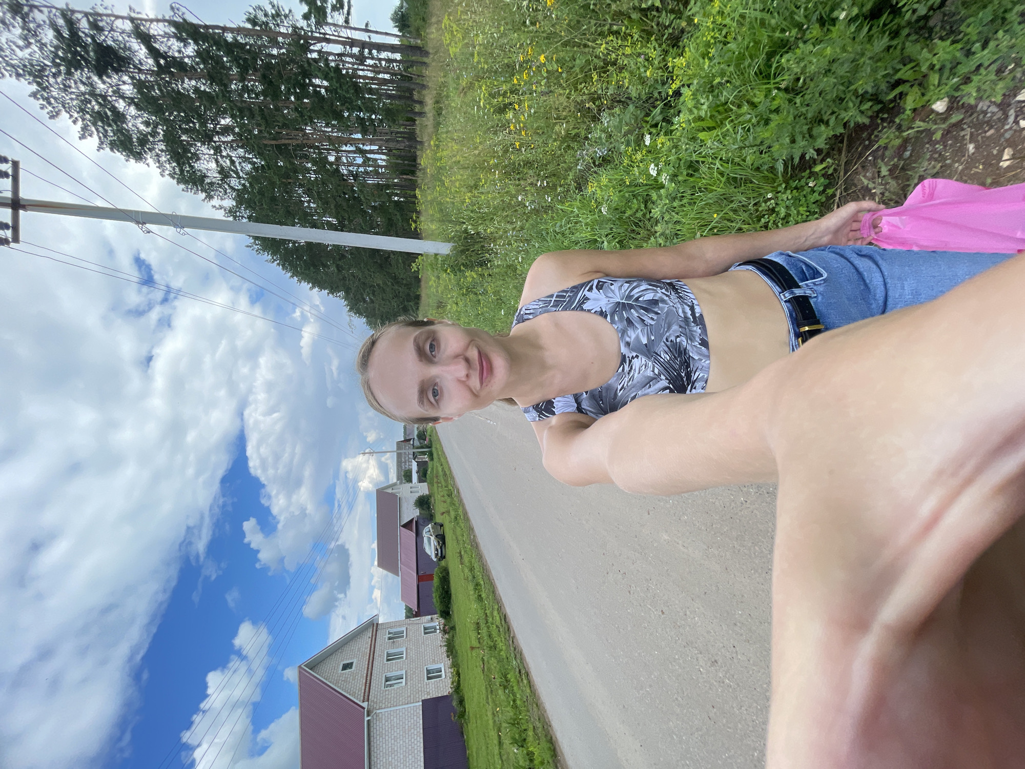A woman with blonde hair pulled back is taking a selfie outdoors on a gravel road in the village of Podshivalovo, Russia. She is wearing a grey and white patterned sports bra and blue jeans with a black belt. She is holding a pink plastic bag in her right hand. The road is lined with green grass and wild flowers on the right and a few houses and trees on the left. In the distance, there is a power pole with wires running across the frame. The sky is blue with scattered white clouds.