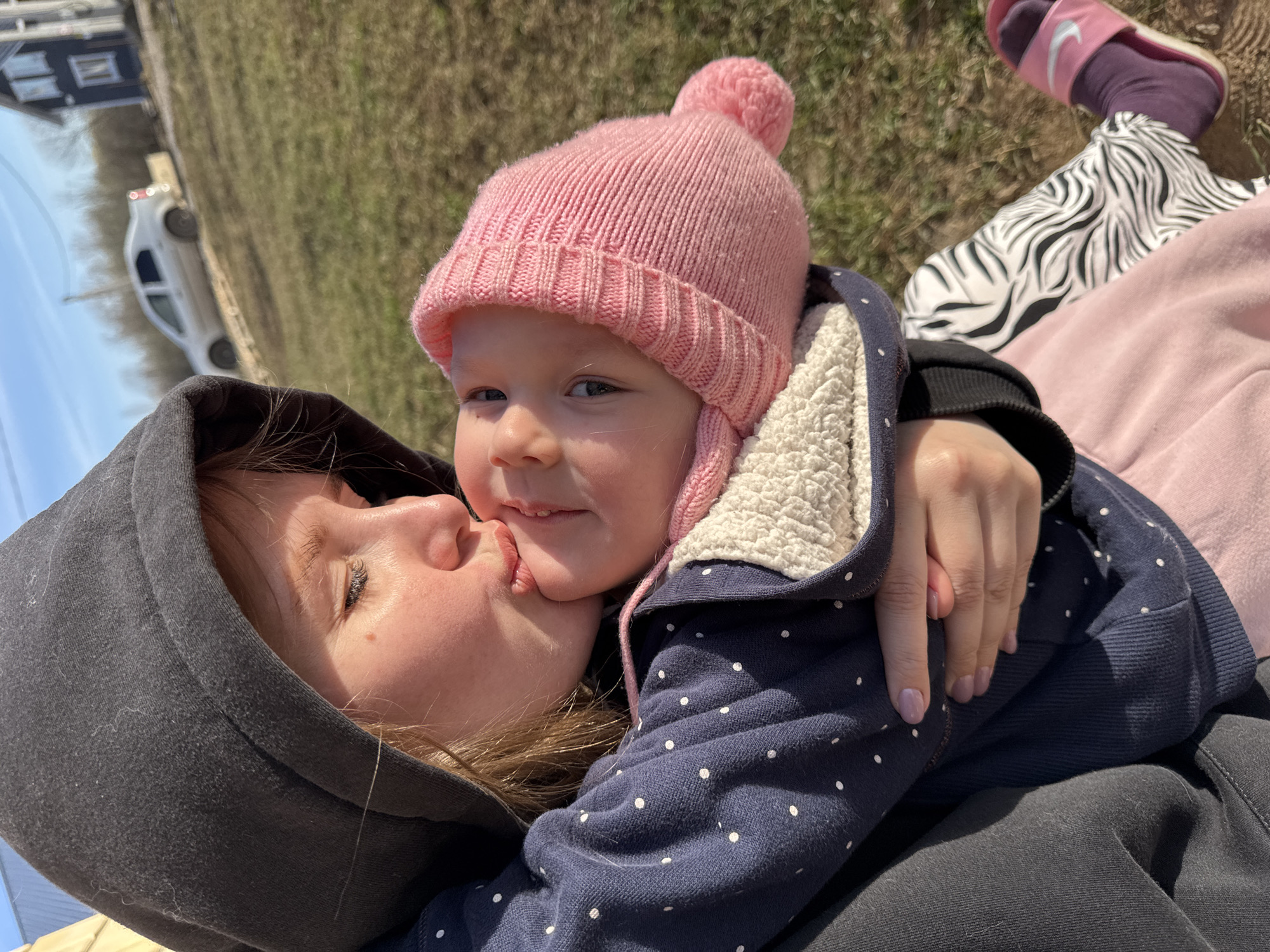 An outdoor, angled close-up photo captures an affectionate moment between an adult and a young child, likely a parent and child. The adult, wearing a dark grey hooded sweatshirt, is kissing the child on the cheek/mouth. The child, in a pink knitted hat with a pom-pom and a navy blue polka-dotted jacket with cream sherpa lining, looks directly towards the camera with a slight smile. The adult's arm is wrapped around the child, and a hand with light-colored nail polish is visible.
The scene is brightly lit by natural sunlight, indicating daytime. The background shows dry, grassy ground, with a clear blue sky visible in the upper left. A white car is parked on what appears to be a dirt road, and a building is faintly visible in the distance, suggesting a rural or suburban setting in Gornyi (Dvurechenskoe S/P), Russia. Another notable detail is a person's foot, possibly belonging to the adult or another individual, wearing a purple sock, a pink Nike slide (with the "Nike" logo clearly visible), and a zebra-print slipper/shoe, resting on a pink garment. The overall emotion conveyed is one of warmth, tenderness, and familial bond.