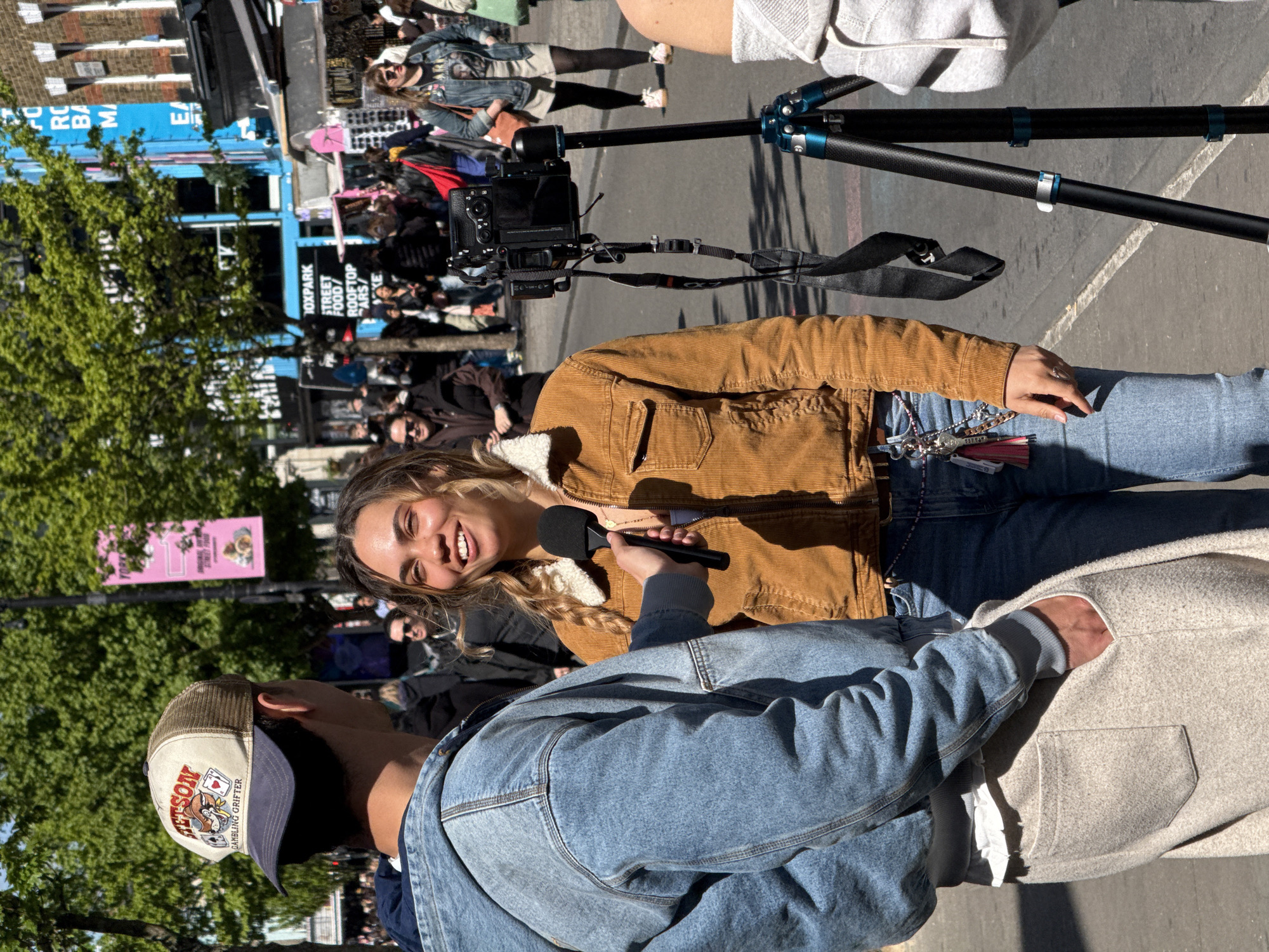 Here is a detailed description of the image:
**General Description:**
The image captures an outdoor scene, likely a street interview. The focus is on a woman being interviewed by a person holding a microphone. The background shows a crowd of people, buildings, and signage. The image appears to be shot during the day.
**People:**
* **Interviewee:** A young woman with long brown hair, wearing a brown jacket, and jeans. She is smiling and looking towards the person holding the microphone.
* **Interviewer:** Only the back of the person interviewing the woman is visible. They are wearing a denim jacket and a baseball cap with an image on it and the word "JETSON." They are holding a black microphone.
* **Background Crowd:** A large group of people is visible in the background, likely pedestrians.
**Objects:**
* **Microphone:** A black microphone is held by the interviewer and pointed towards the interviewee.
* **Camera on Tripod:** A camera is set up on a tripod, pointed towards the woman being interviewed.
* **Signage:** Signs with text are visible in the background, including one that says "DOXPARK STREET FOOD/ ROOFTOP BARS/ ."
* **Buildings:** Buildings are visible in the background.
**Actions and Interactions:**
* **Interview:** The primary action is an interview taking place between the woman and the person holding the microphone.
* **Filming:** A camera is set up to record the interview.
* **Crowding:** People are gathered in the background.
**Environment:**
* **Outdoor Setting:** The scene is outdoors, likely on a street or sidewalk.
* **Daylight:** It appears to be daytime, with bright sunlight.
**Location (Based on Signage):**
*The sign reads "DOXPARK STREET FOOD/ ROOFTOP BARS/ ". "DOXPARK" is located in the city of London, United Kingdom.
**Additional details**
* The interviewer is wearing a baseball cap. The text says "JETSON GAMBLING GRIFTER".
* The woman is wearing jeans and has a keychain with multiple elements hanging from her jeans.
* A grey coat is being held by the interviewer.