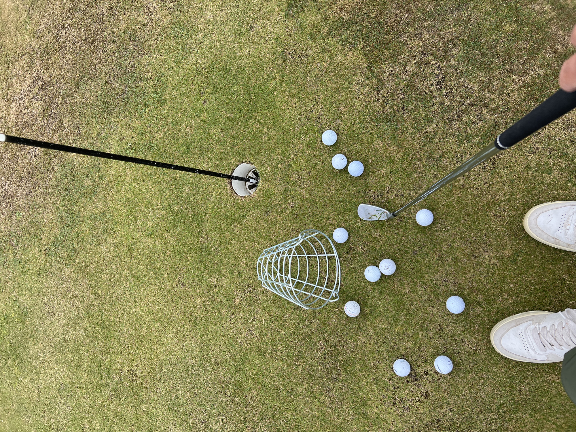 An overhead shot shows a person on a golf course in Kawle Górne, Poland, practicing their putting. Several white golf balls are scattered across the green, with a wire basket typically used for collecting practice balls positioned near them. A golf flagstick stands upright in the hole. The grass appears somewhat dry, with patches of brown visible. The perspective suggests the person is standing and holding a golf club, preparing to strike a ball.