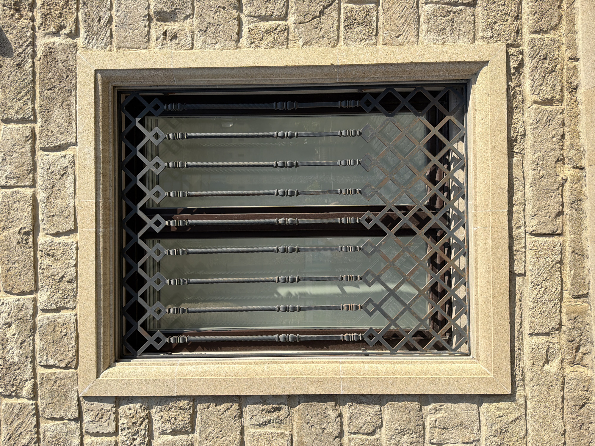 This is a frontal, eye-level, medium shot of a window with a decorative iron grate set into a rough, light brown stone wall in Sabail Raion, Azerbaijan. The window is rectangular with a beige stone frame that has a beveled edge. The iron grate is black and features a diamond pattern at the bottom, with vertical twisted bars extending upwards. The glass behind the grate is opaque and reflects some light, suggesting it may be frosted or have curtains. The stone wall is constructed from irregular-shaped blocks of varying sizes, with visible mortar lines. The lighting suggests it is daytime, with shadows indicating sunlight. No people or animals are present in the image.