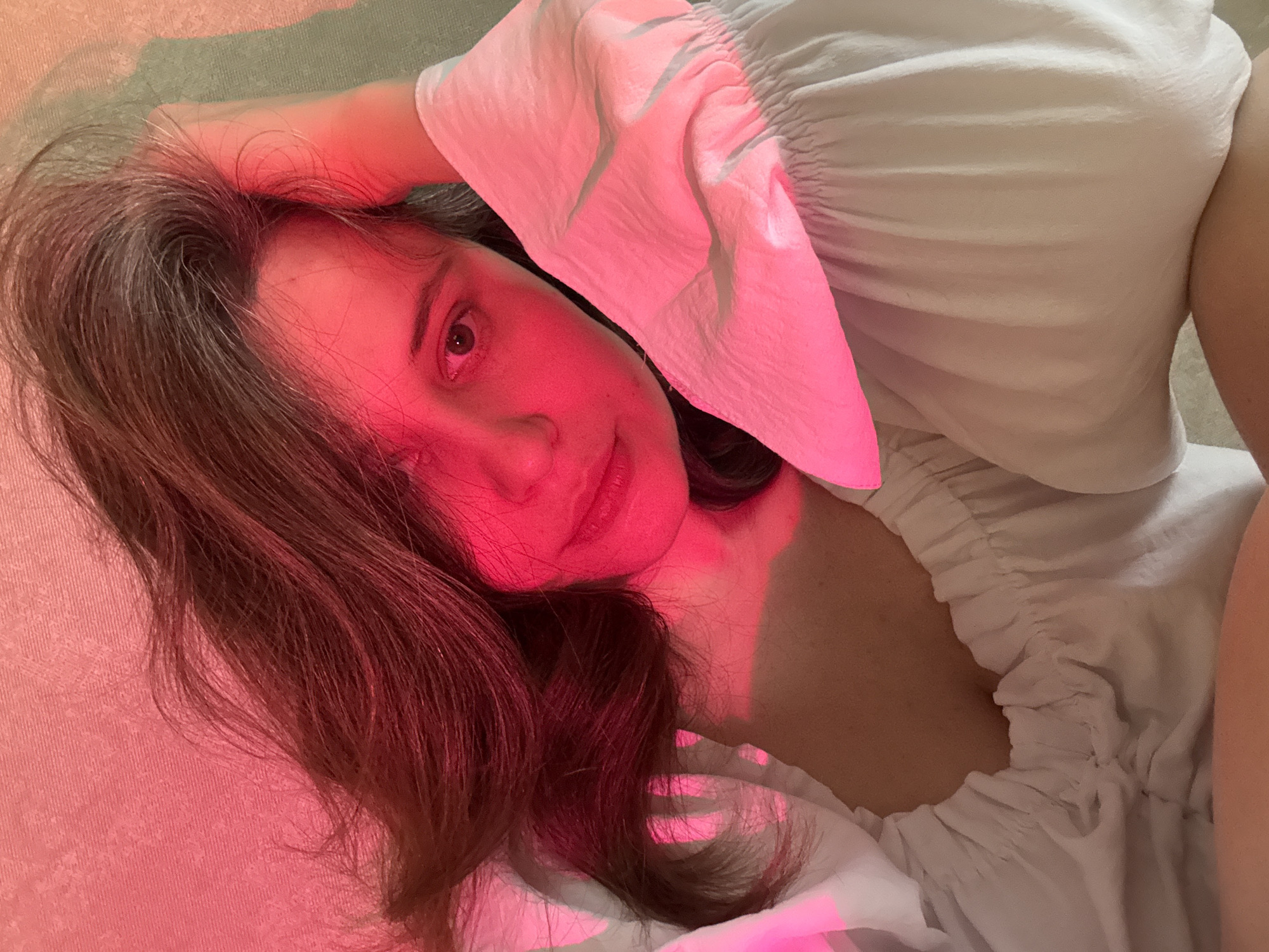 A woman is lying on her back. She has long brown hair and is wearing a white top. She is in a room illuminated by a red light source. There is no evidence to suggest that the image was taken in the city of Chaikovsky, Russia.