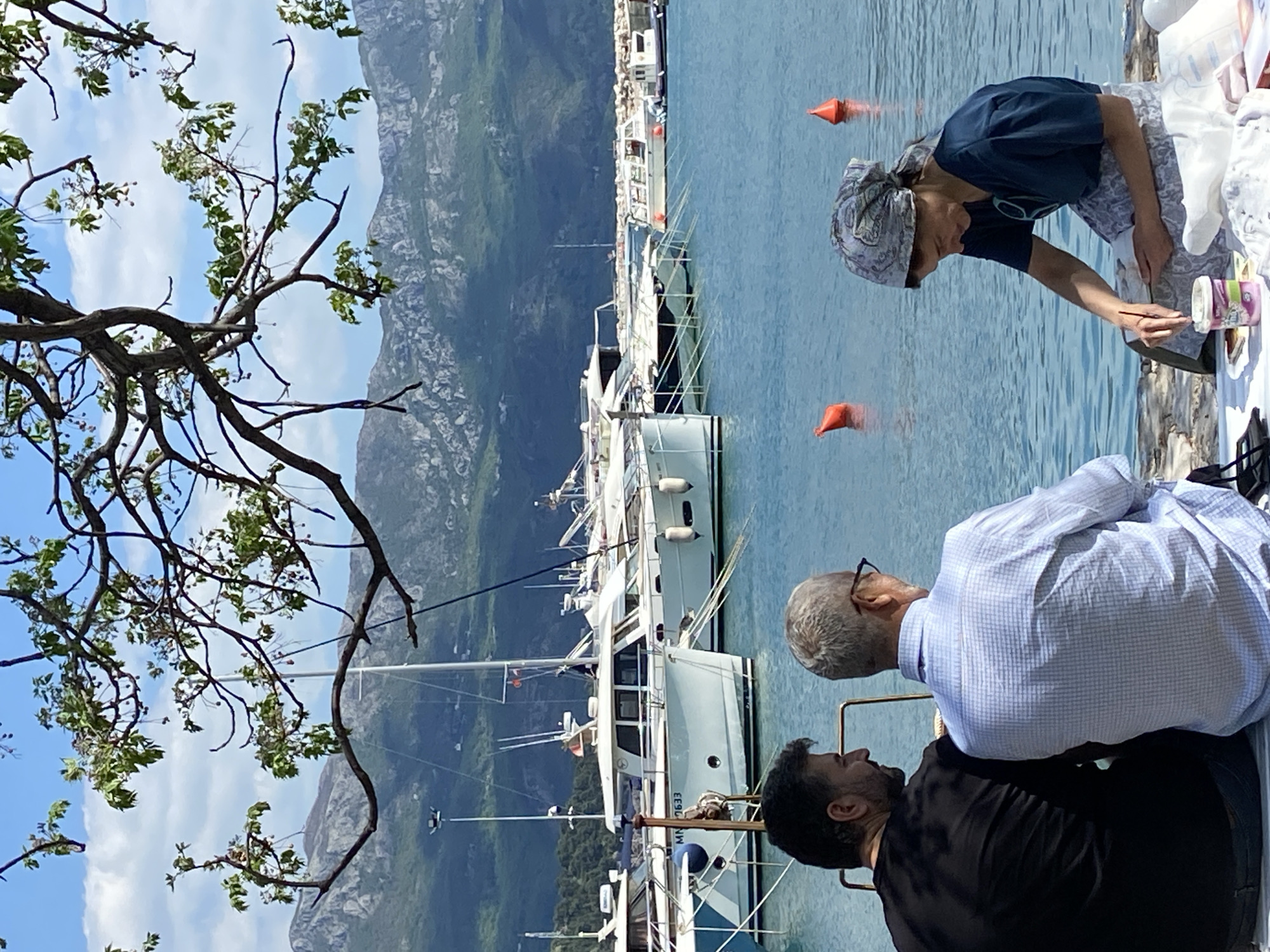 Two men stand beside a seated woman painting on a dock in Budva, Montenegro. Behind them, yachts are moored in the harbor. Mountains rise in the background under a partly cloudy sky, partially obscured by tree branches in the foreground. Two orange buoys are visible in the water. One man is wearing a light blue checked shirt and glasses, while the other is in a black shirt. The woman wears a dark blue shirt and headscarf.