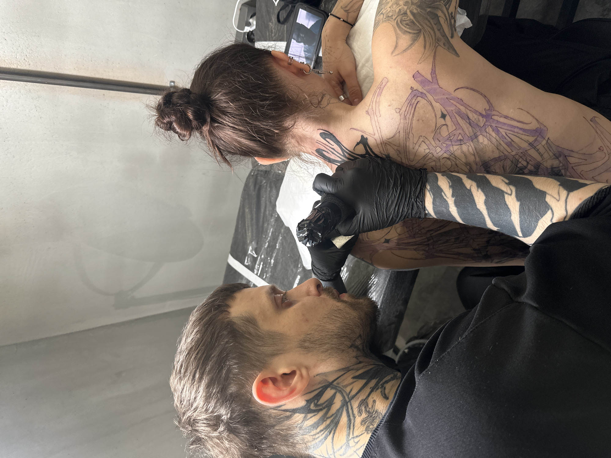 A man with a beard and neck tattoos is tattooing the back of a woman lying on a table. The man is wearing black gloves and using a tattoo machine. The woman has a bun and is lying face down with a phone visible above her head. The tattoo being applied is a large design in purple and black. The scene appears to be taking place in a tattoo studio, and I lack information to determine if it is Moscow, Russia.