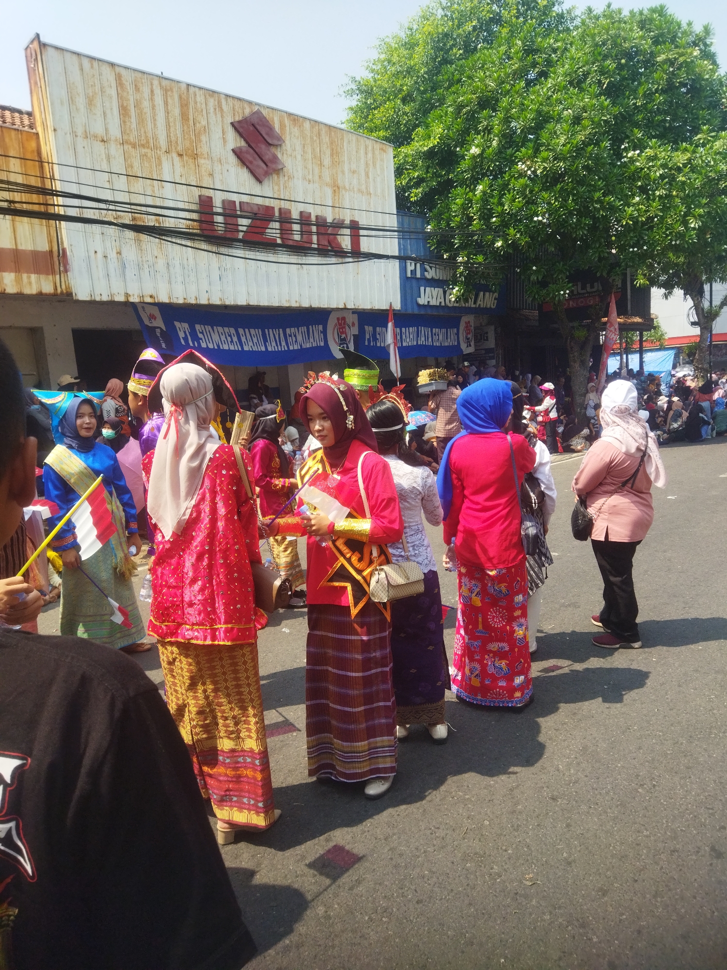 In the city of Wonogiri, Indonesia, a group of people are gathered on a street, with a Suzuki dealership and other businesses visible in the background. Several individuals, predominantly women, are dressed in traditional attire. Some wear colorful patterned sarongs and long-sleeved tops, while others are adorned with headscarves in vibrant colors like red, blue, and white. One woman in a red outfit holds a bundle of what appear to be golden fan-like objects. Another woman, also in red, is holding a small white flag with red accents on a stick. In the foreground, the back of a person wearing a black t-shirt is visible. The street is paved, and there are shadows cast on the ground, indicating a bright, sunny day. A large, leafy green tree stands to the right of the Suzuki building. The overall scene suggests a cultural event or parade taking place on the street.