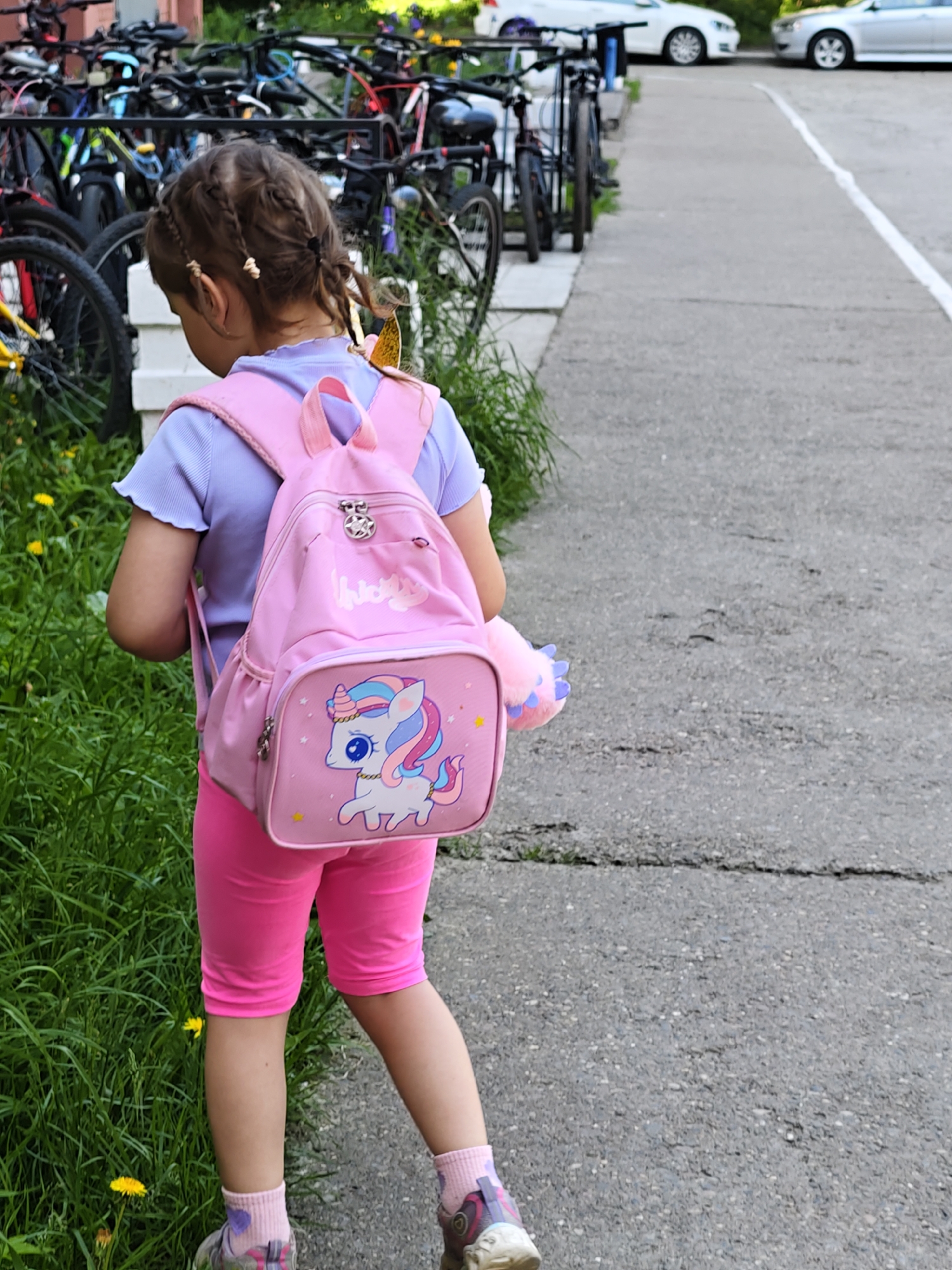A young girl with braided hair, wearing a light purple t-shirt and bright pink shorts, walks away from the camera on a paved path. She has a pink backpack with a unicorn design on it, and a pink stuffed animal attached to the strap. The path is bordered by a grassy area with yellow flowers on the left and parked bicycles on the background. Two white cars are parked in the distance. The scene appears to be outdoors in a residential area, possibly in Chaikovsky, Russia, based on the context provided.