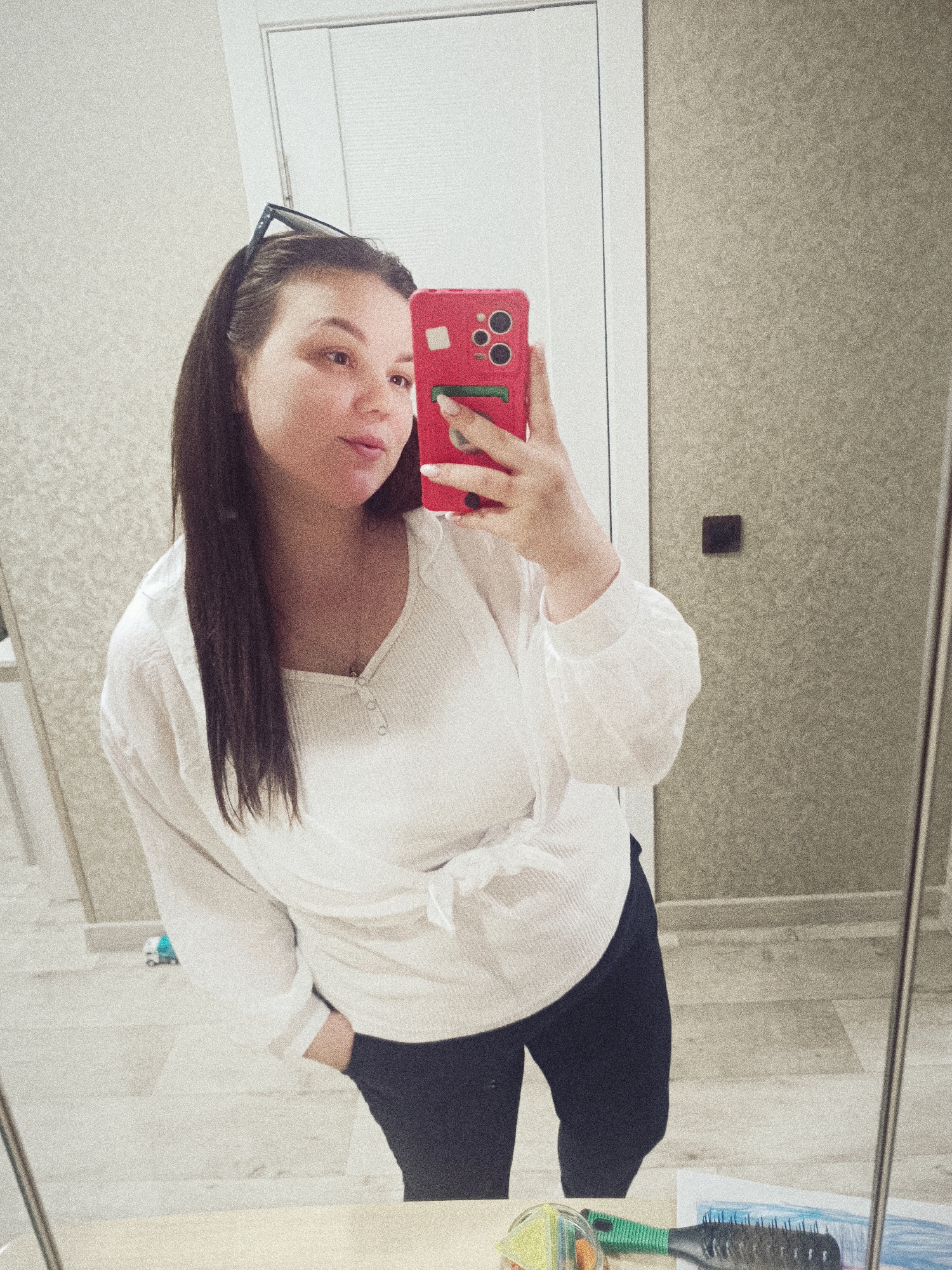 A woman is taking a selfie in a bathroom. She is wearing a white shirt tied at the waist and dark pants. Sunglasses are perched on her head. She is holding a red phone in her right hand. The bathroom has a white door and a beige wall. A mirror reflects the scene. In the bottom of the frame, there are art supplies and a hairbrush on a surface. Based on the instructions and the name in the prompts, the location is Прикамский, Russia.