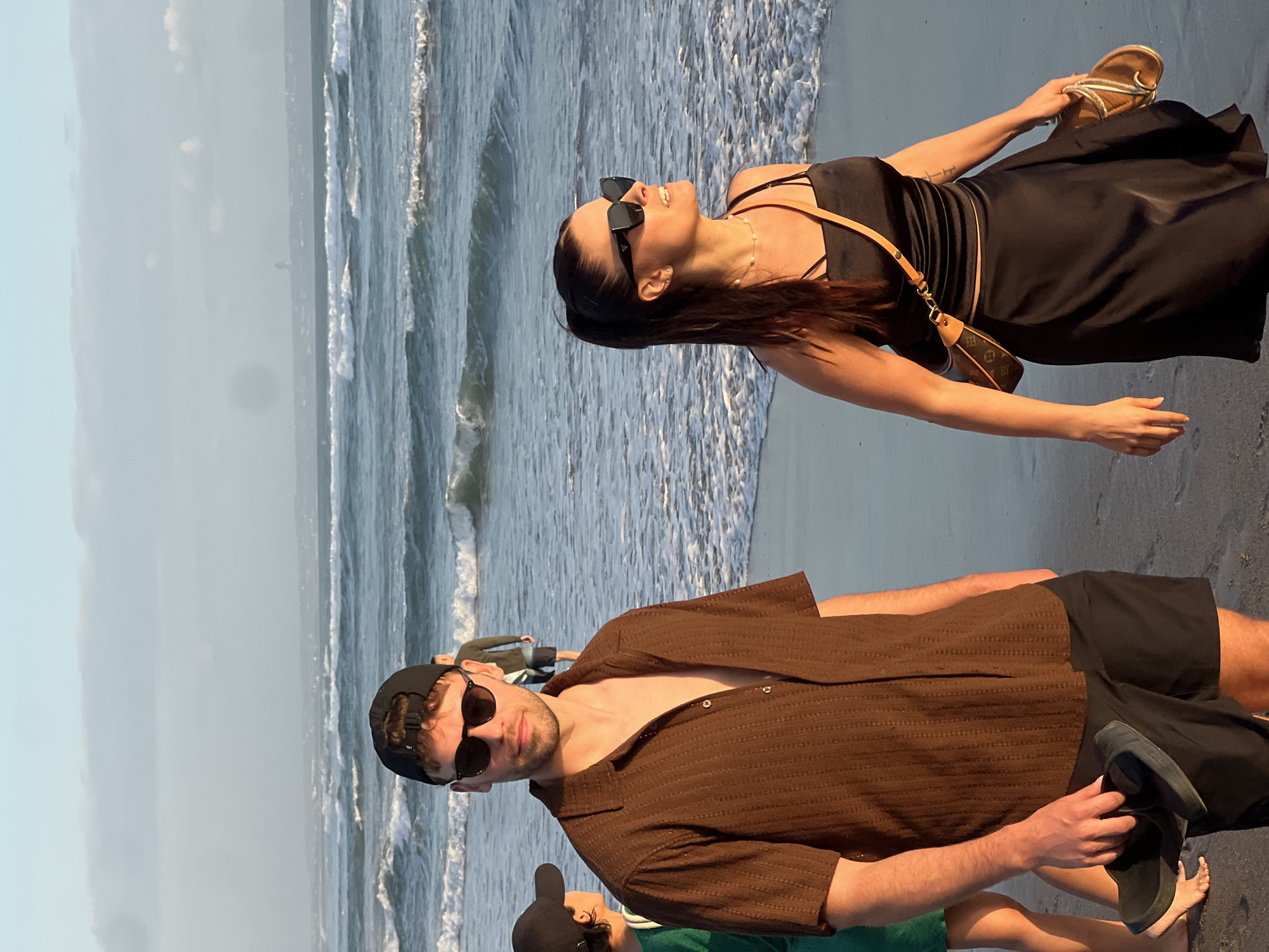 Two people, a man and a woman, are walking on the beach in North Kuta, Indonesia, during sunset. The man, in the foreground on the left, wears a brown short-sleeved button-up shirt, black shorts, a black baseball cap, and sunglasses. He is holding a pair of black sandals in his right hand. The woman, to his right, wears a black slip dress, sunglasses, and a pearl necklace. She carries a small brown and tan shoulder bag and a pair of gold sandals in her right hand. The beach is sandy, with waves breaking in the background. The sky is a soft blue with some clouds, and the setting sun casts a warm glow on the scene. Other beachgoers are visible in the distance.