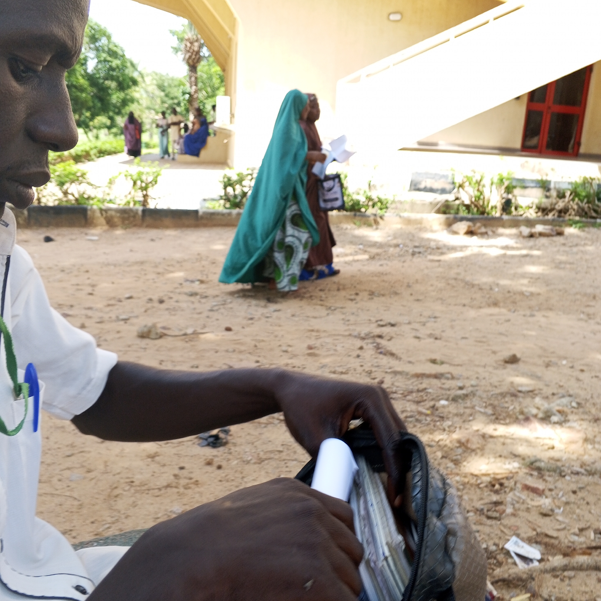 In Arawa, Nigeria, a close-up shot focuses on the hands of a person wearing a white shirt and dark skin, sorting through a dark bag containing several rectangular items, possibly currency or documents. The background shows a sandy ground with scattered debris and vegetation, under the shade of an overhang. In the middle distance, three individuals, two women in colorful Islamic attire, walk away from the camera, one holding papers. Further in the background, a building with a red-framed window is visible. The lighting suggests it is daytime.