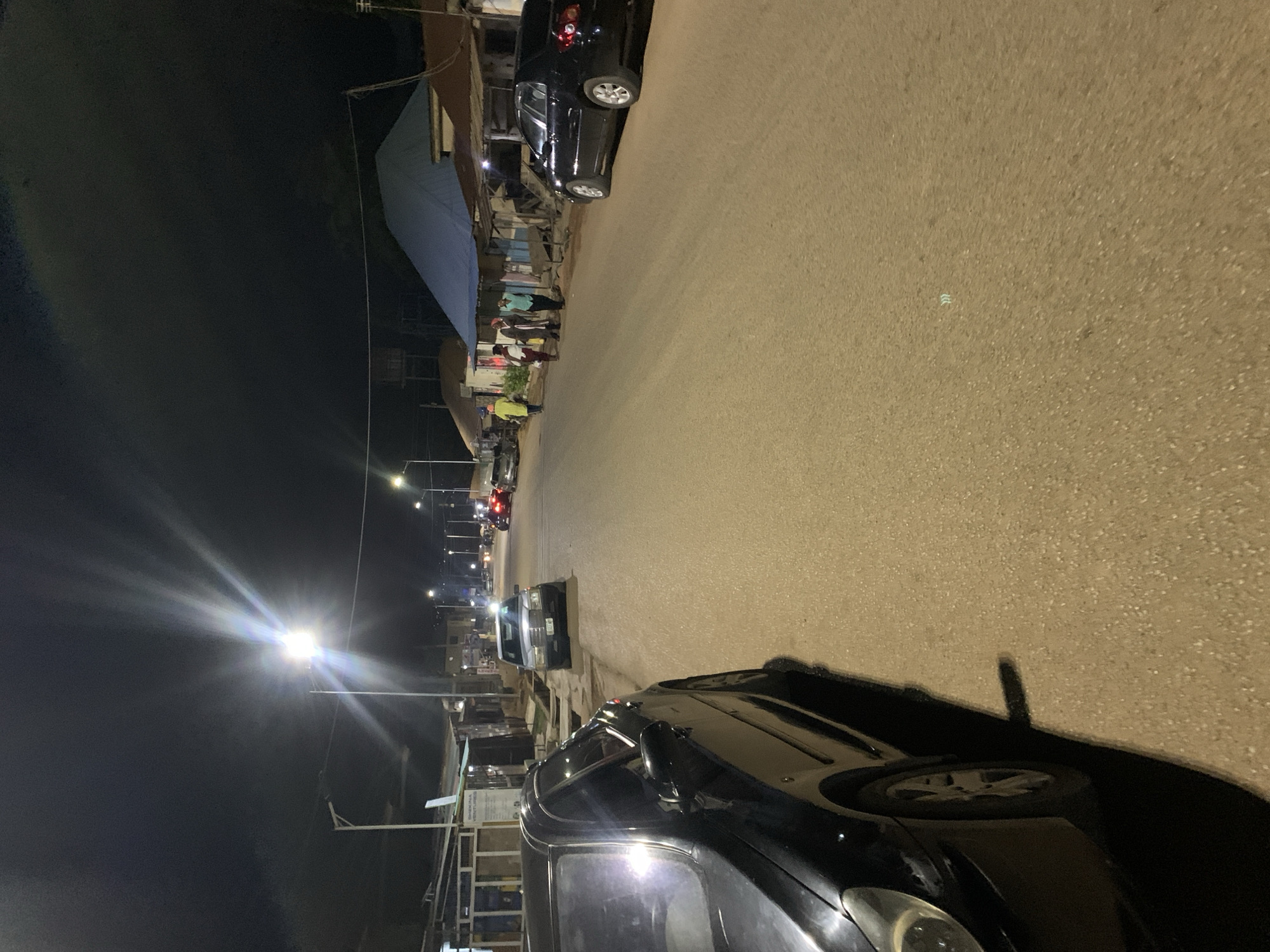 The image depicts a street scene at night in Kabong, Nigeria. The street is paved and illuminated by distant streetlights. Parked cars line both sides of the street. A group of people are gathered on the left side of the road, near buildings. The environment is dark, with artificial light sources providing visibility.