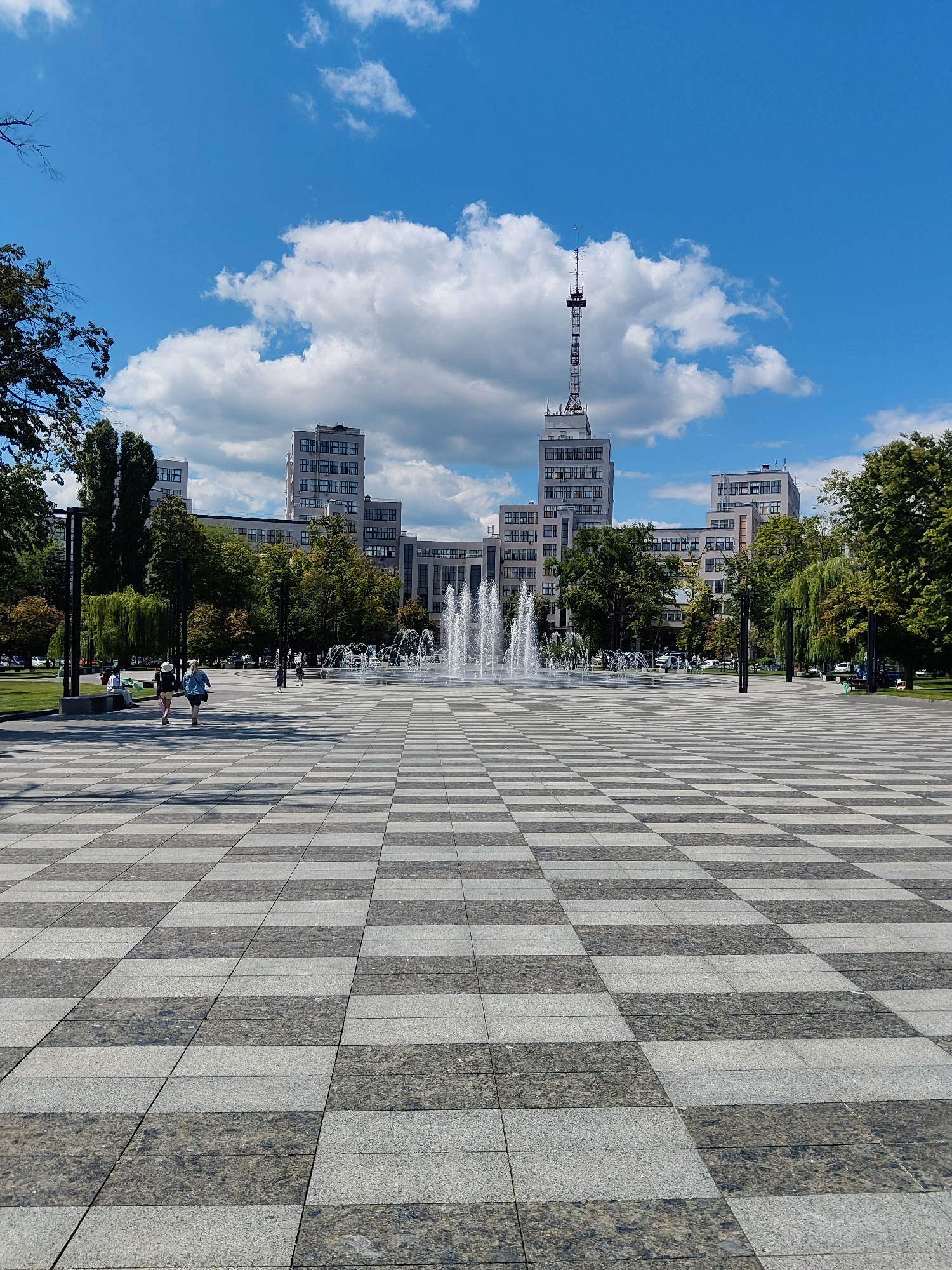 This image captures a wide-angle view of Freedom Square in Kharkiv, Ukraine, on a sunny day with a bright blue sky and fluffy white clouds. The foreground is dominated by a large, checkered plaza paved with alternating dark gray and lighter gray granite tiles. In the mid-ground, a fountain sprays water upwards, creating a misty effect. Behind the fountain, a strip of green grass is visible, lined with trees. Beyond the trees, several large, multi-story buildings are situated. The most prominent building in the center features a tall communication tower on its roof. Several people are walking and sitting in the plaza and on benches in the background. The overall scene depicts a public urban space with architectural elements, natural features, and human activity.