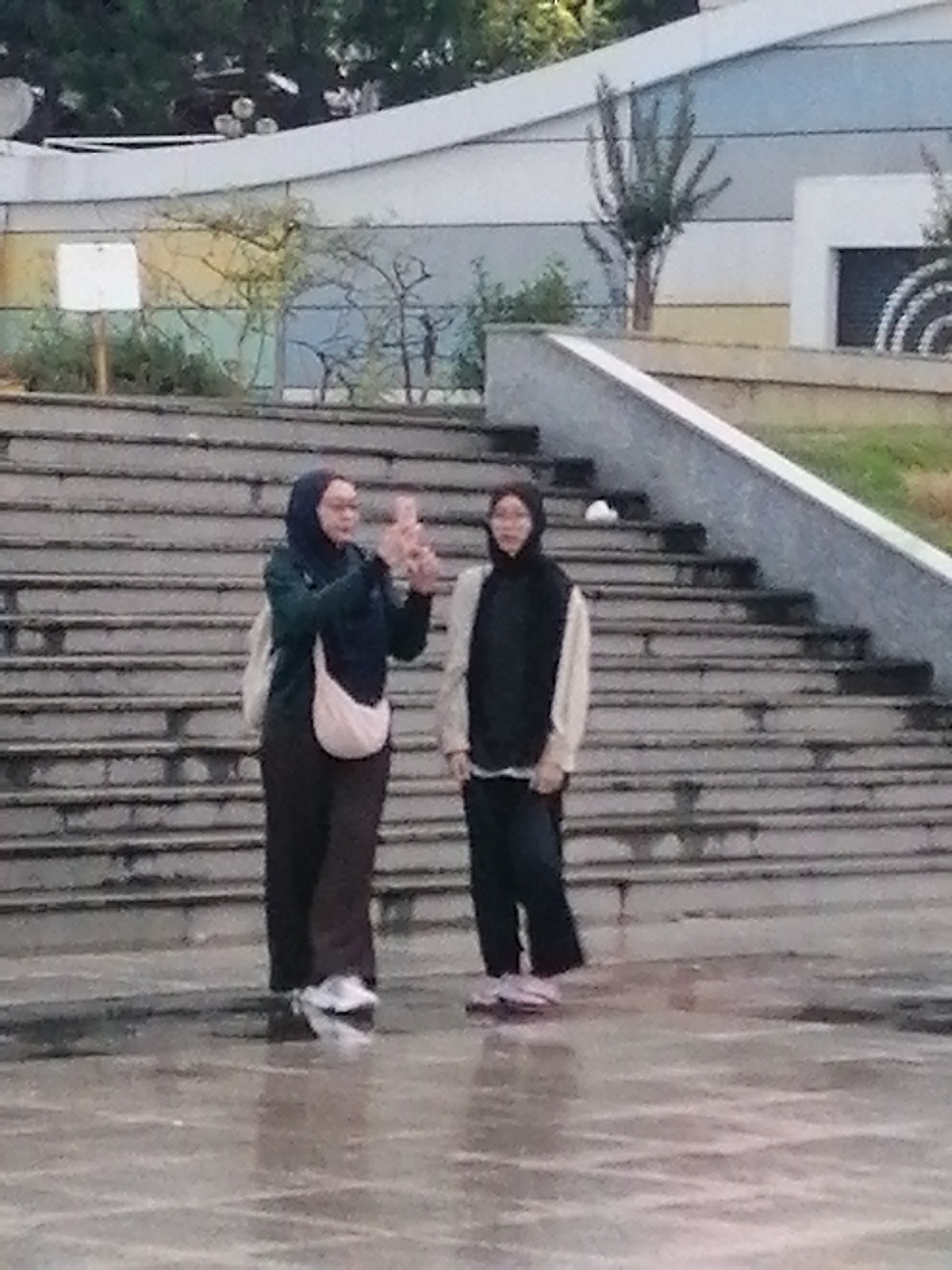 Two individuals, likely women, are standing on a wet, paved surface in front of a set of concrete stairs. The individual on the left, wearing a dark hijab, dark pants, and a light-colored bag slung across her chest, is holding up a pink object, possibly a phone, as if taking a picture or video. The individual on the right, also wearing dark pants and a light-colored top with a dark vest over it, is looking towards the left. The stairs ascend to a modern-looking building with horizontal stripes in shades of blue, white, and yellow. Trees and bushes are visible behind the building. The ground appears to be wet and reflective, suggesting it may have recently rained. The scene is set outdoors in what could be a public plaza or park area. This setting does not appear to be within the city of Da Lat, Vietnam.