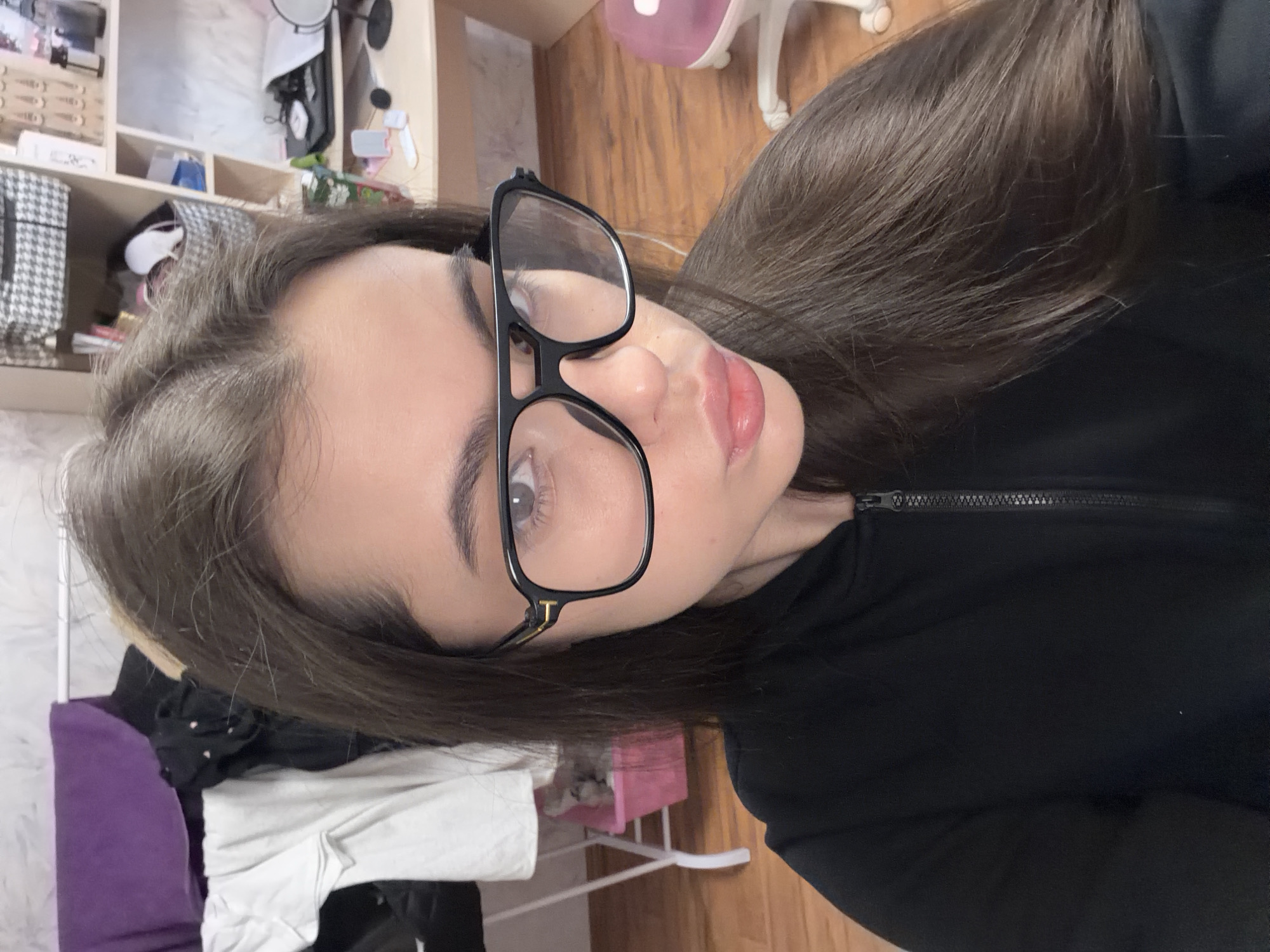 The image is a selfie of a young woman with long, dark hair and light eyes. She is wearing black-framed glasses and a black jacket. The background shows a room with a shelf containing various items, including a mirror and storage boxes. There is also a chair visible. Based on the presence of Cyrillic text on some of the items on the shelf (visible at the top left of the image), the location may be Chaikovsky, Russia.