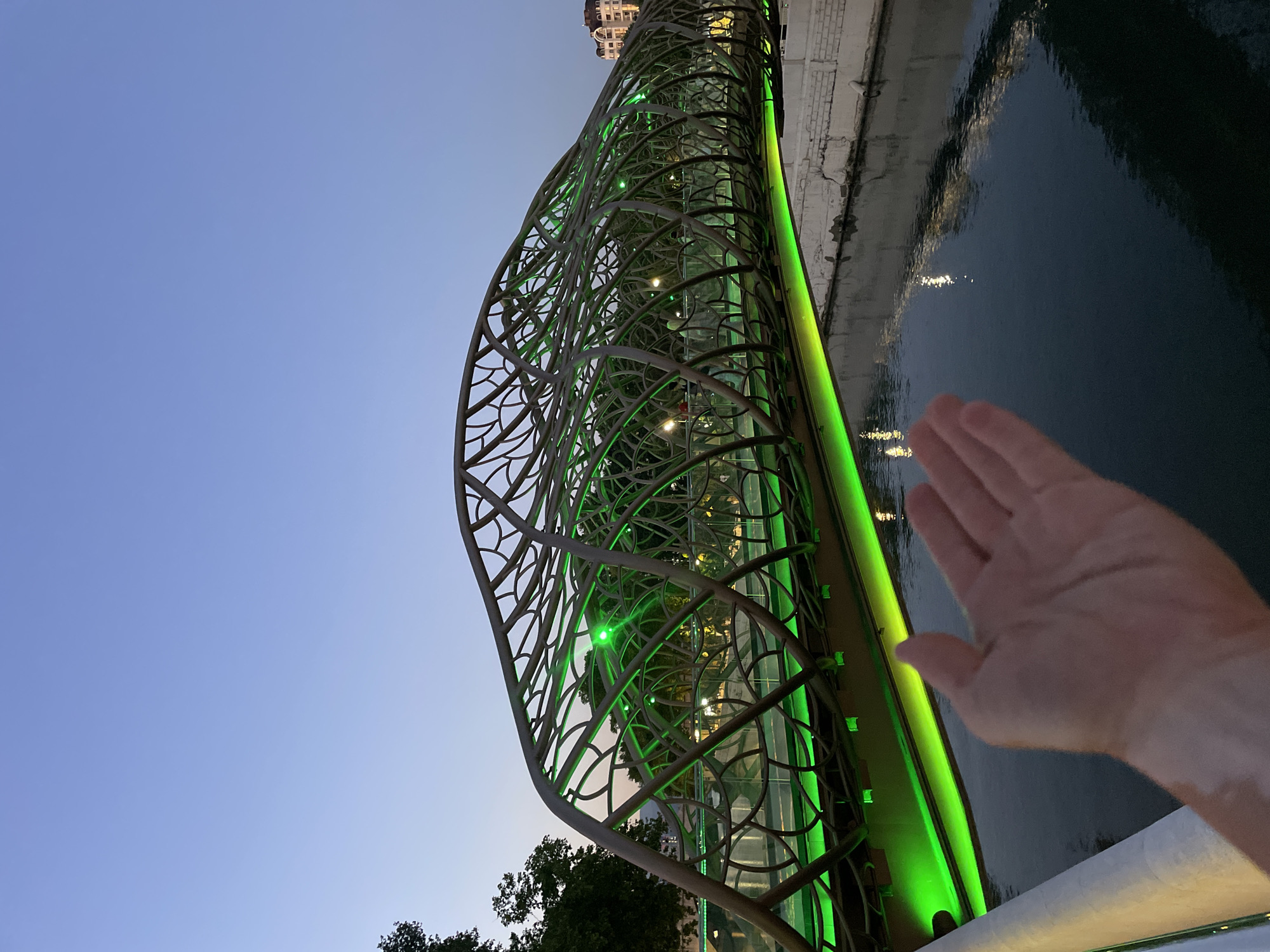This is a photograph taken at twilight on the Starfall Bridge (also known as the Bridge of Fairy Tales) in Sochi, Russia. The bridge, a pedestrian crossing over the Matsesta River, is illuminated with vibrant green LED lights that highlight its intricate, branch-like metallic structure. The bridge's organic, undulating form is reminiscent of a cocoon or a natural formation.
In the foreground, a person's left hand is extended towards the camera, palm open. The hand exhibits patches of vitiligo. The hand is positioned to the left of the image, with the rest of the bridge and river occupying the frame.
The water of the Matsesta River below reflects the green illumination from the bridge, creating a shimmering effect. The riverbank on the right is lined with a white concrete wall, showing some signs of age and wear. In the background, beyond the bridge, trees and the facades of buildings are visible, illuminated by the fading light and the bridge's glow. The sky is a deep, clear blue, transitioning from the day's end. The overall atmosphere is serene and visually striking, emphasizing the modern architectural design of the bridge against the natural setting.