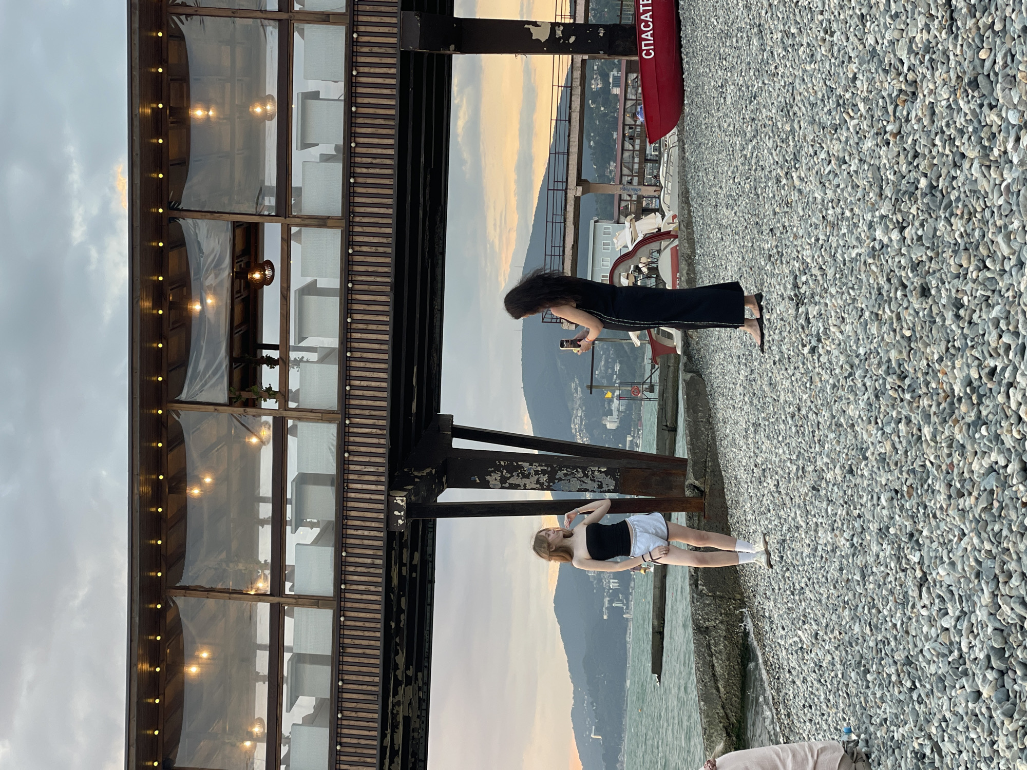 Two young women are on a pebble beach in Sochi, Russia, with a body of water to their left and a pier structure with outdoor seating above them. The woman on the left, with blonde hair, wears a black strapless top and light grey shorts, standing near the water's edge. The woman on the right, with dark hair, is dressed in a long black dress and is taking a photo with her phone of the woman on the left. Behind them, a red boat with "СПАСАТЕЛ" (Rescuer) written on it is visible, along with other boats and the shoreline with buildings and hills in the distance. The sky is cloudy with hints of sunset colors.