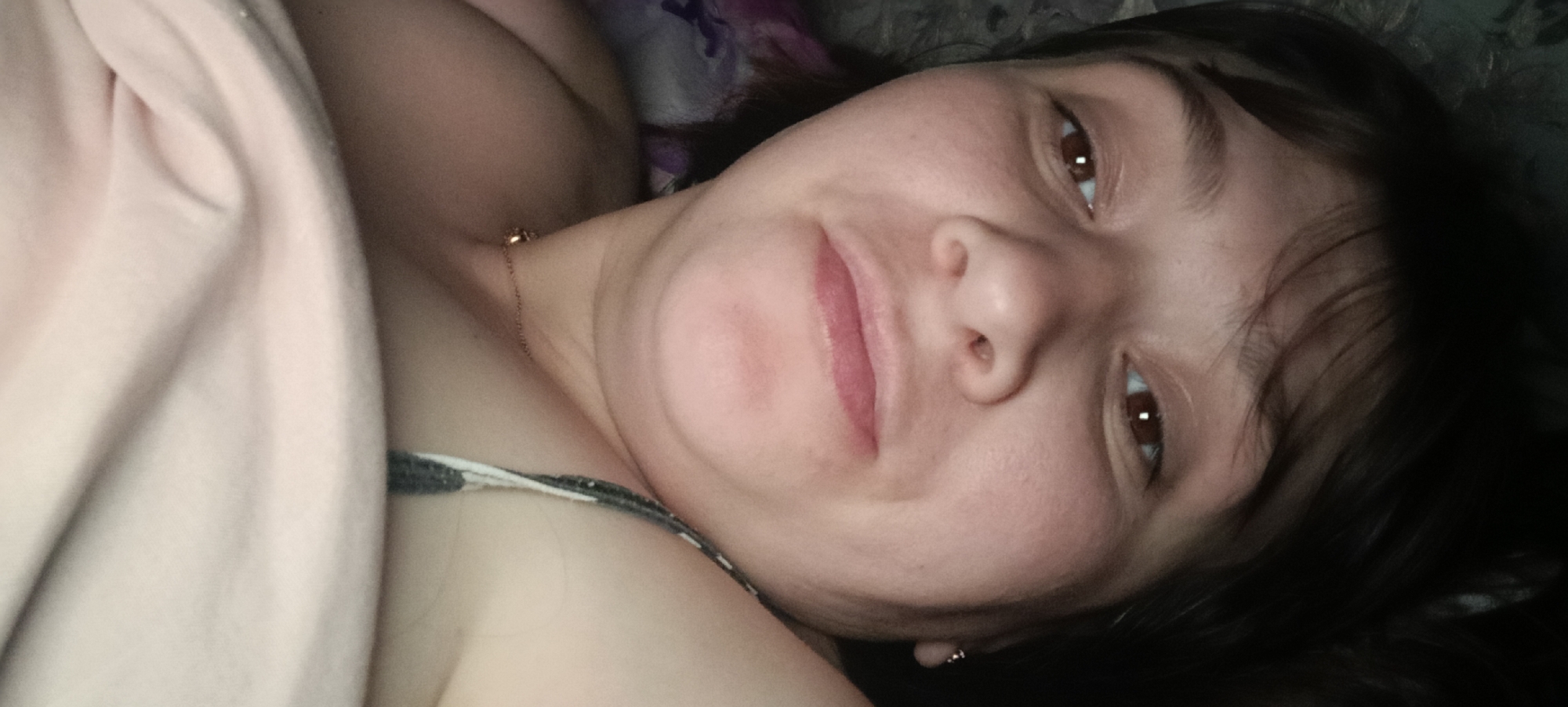 The image is a close-up, low-angle selfie of a young Caucasian woman lying down. She is partially covered by a light pink blanket or sheet, and her dark hair is spread around her head. Her eyes are dark brown and looking towards the camera. She is wearing a patterned bra strap. A thin gold necklace is visible around her neck. The background appears to be dark and textured, possibly a bedspread or wall. There is no indication of the location being in Хохлы, Russia.